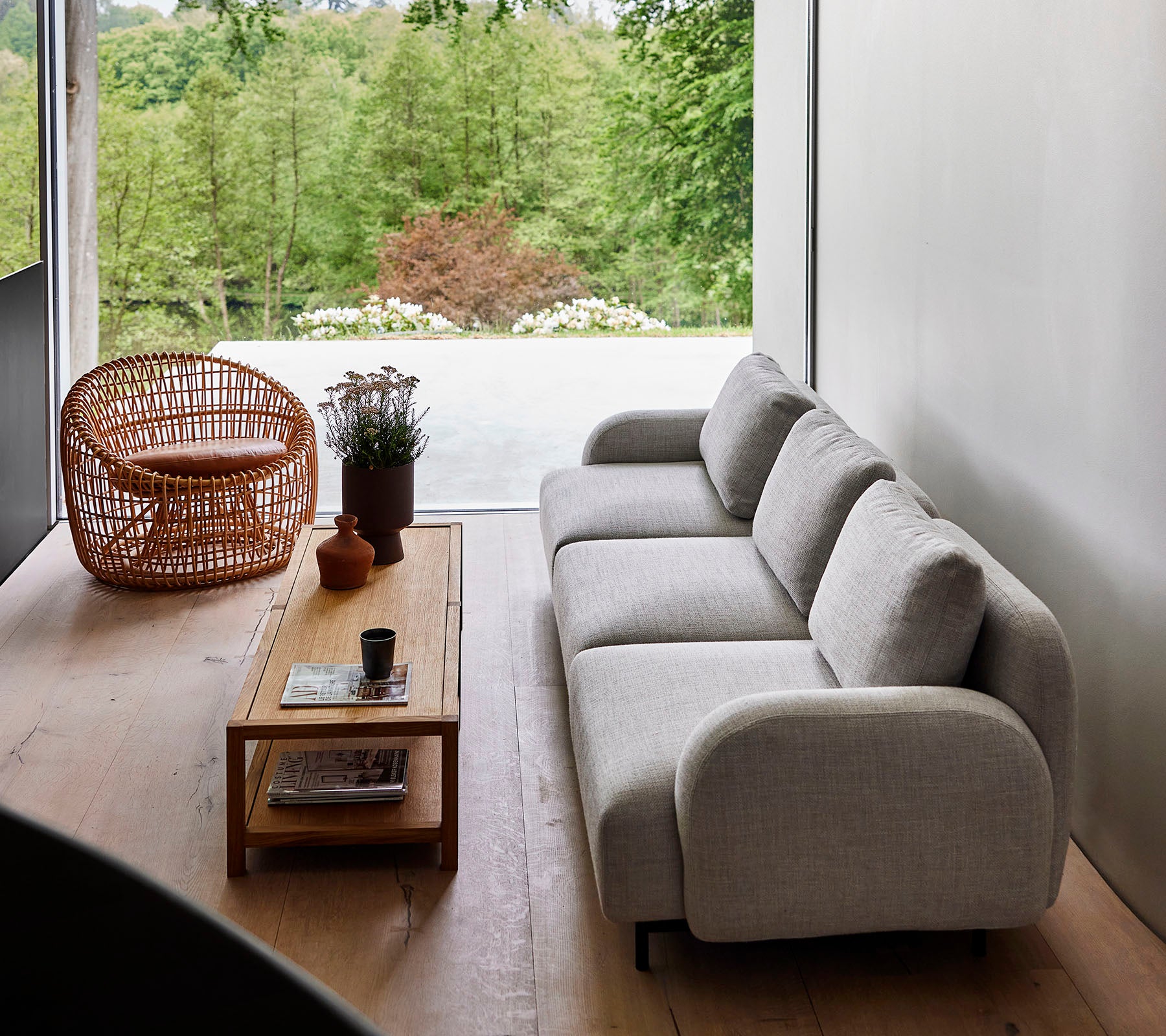 Sofá moderno de tela clara combinado con una mesa de café de madera y una silla de mimbre, rodeado de vegetación. La luz solar natural realza la atmósfera serena.