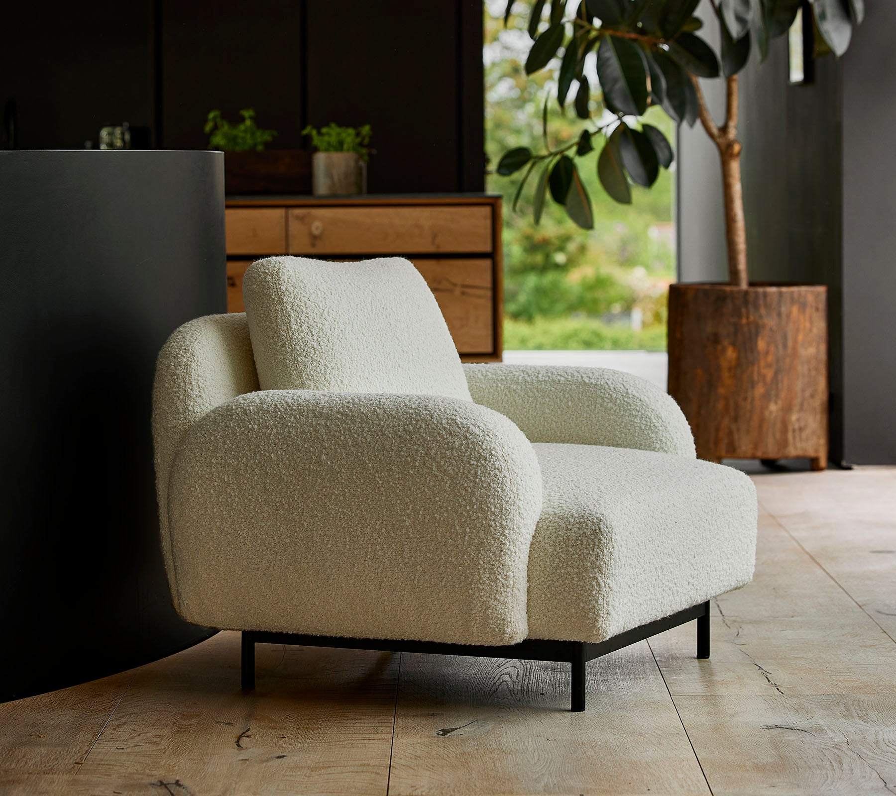 Silla de lounge blanca y acogedora con reposabrazos redondeados y una textura suave, situada frente a un fondo de vegetación y muebles modernos.