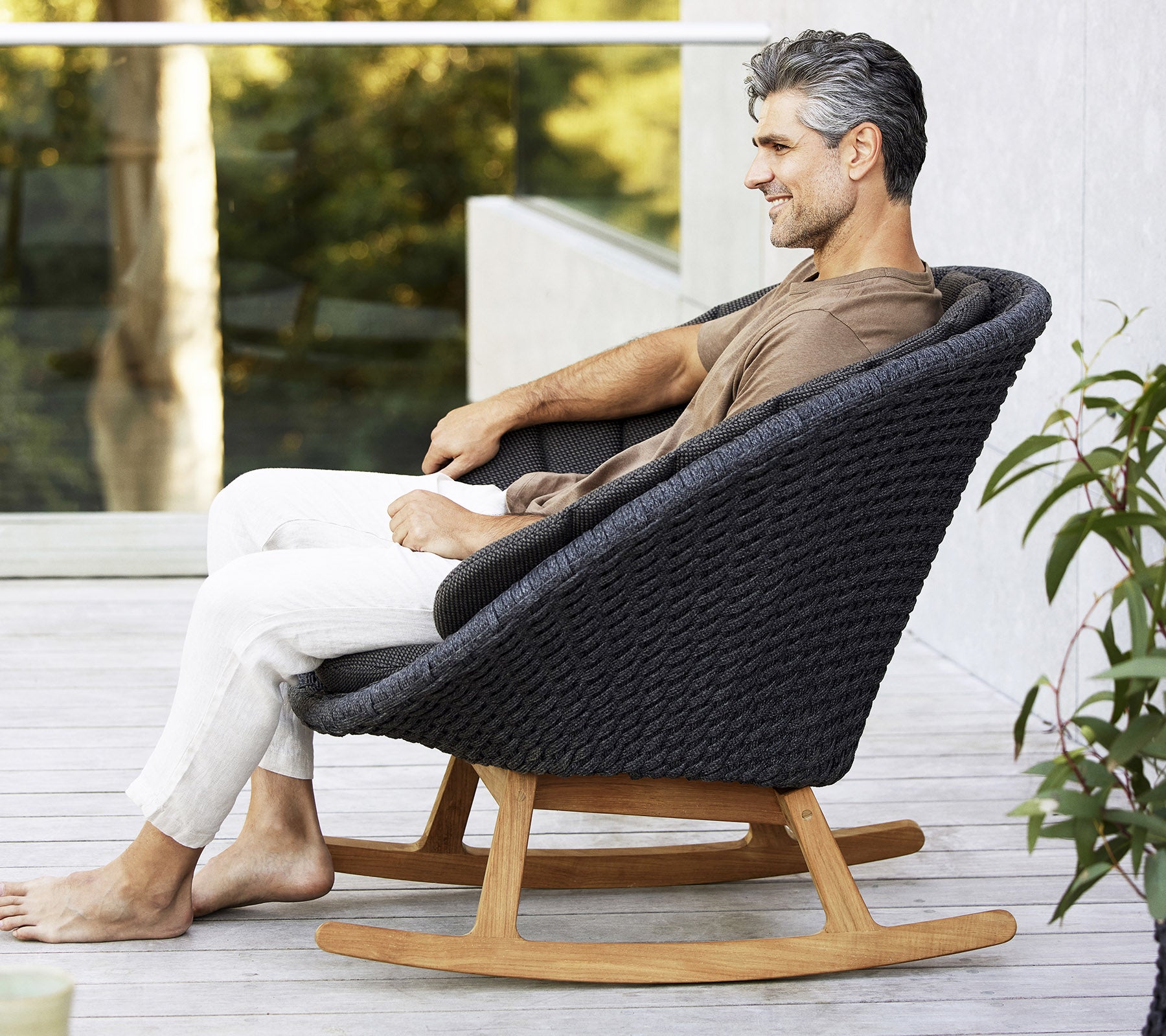 Un hombre se sienta relajado en una silla mecedora moderna y elegante, con un diseño tejido y una base de madera natural, rodeado de vegetación.