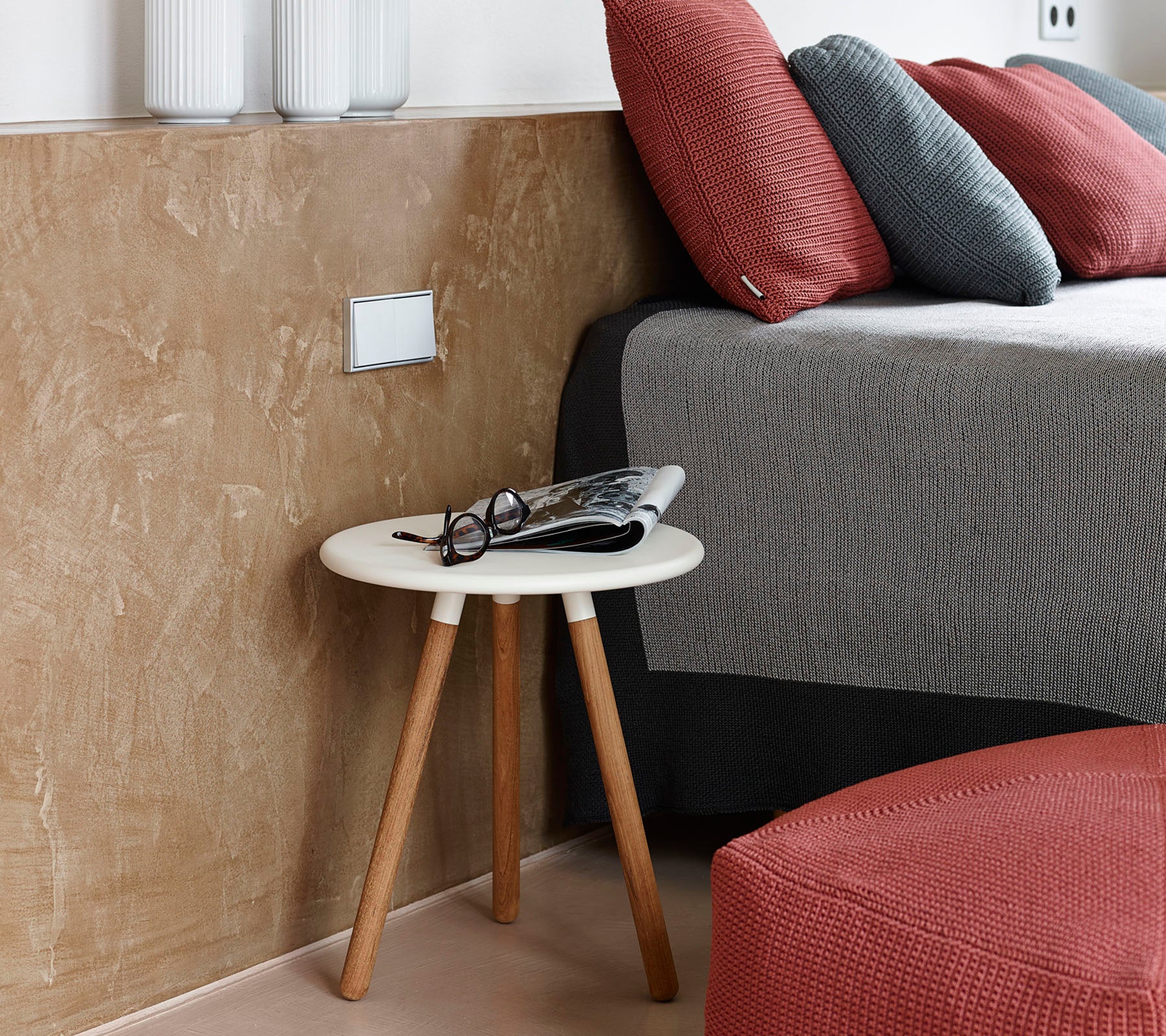 Una escena de dormitorio acogedora con una mesa auxiliar baja con gafas y una revista al lado de una cama bien hecha con almohadas texturizadas.