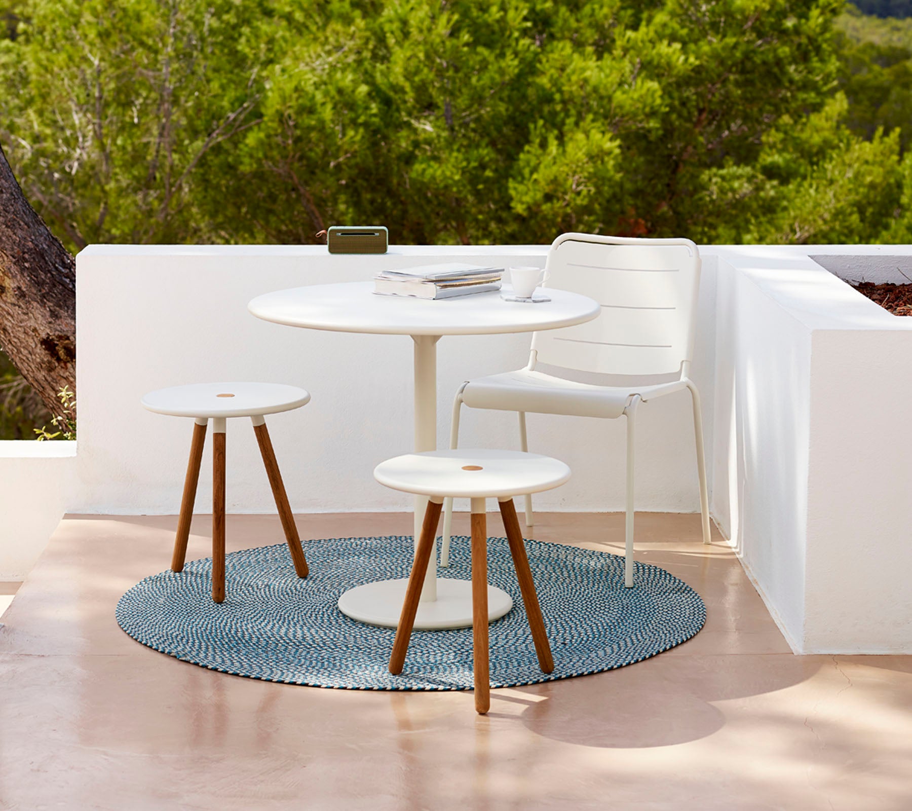 Conjunto de mesa de exterior moderno con una mesa redonda, dos taburetes y una alfombra texturizada, rodeado de vegetación. Ideal para relajarse.