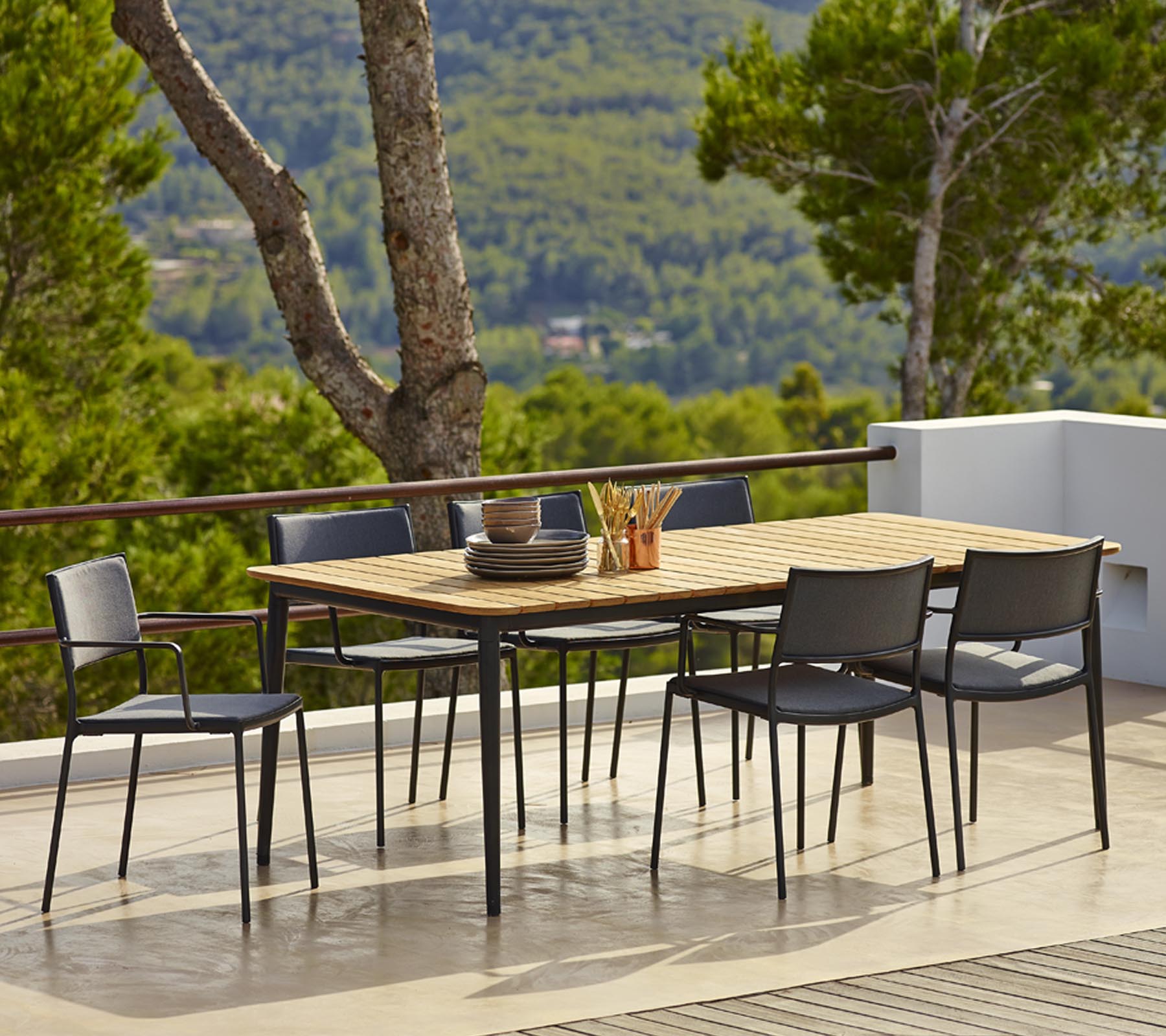 Conjunto de mesa de comedor al aire libre con una mesa de madera rectangular y sillas negras, rodeado de vegetación y vistas escénicas.
