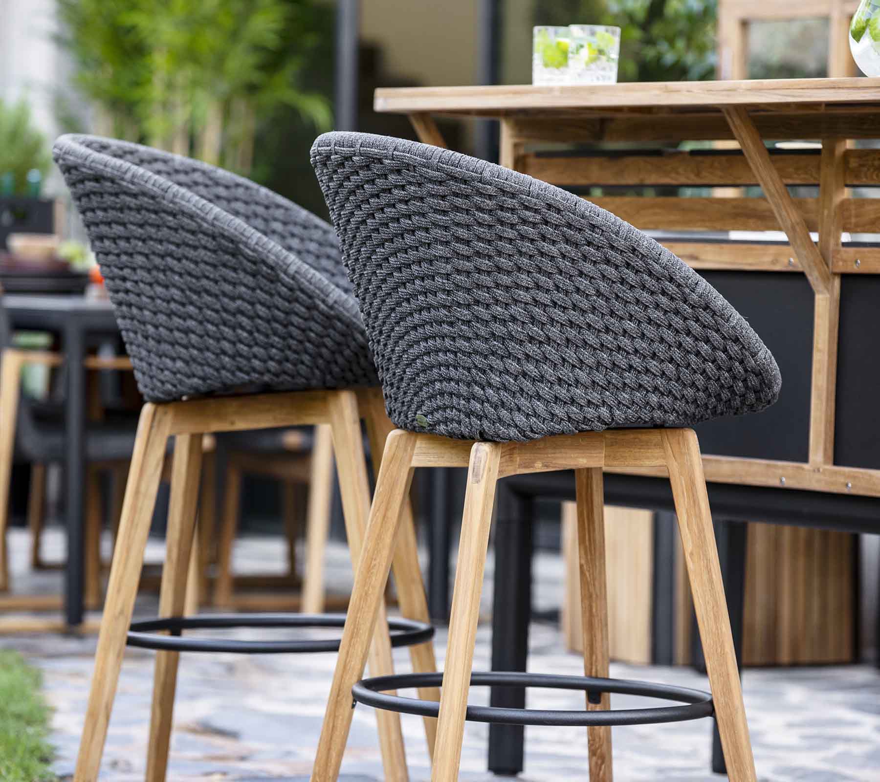 Taburetes de bar elegantes con respaldos grises trenzadas y patas de madera, dispuestos cerca de una mesa de madera en un entorno contemporáneo.