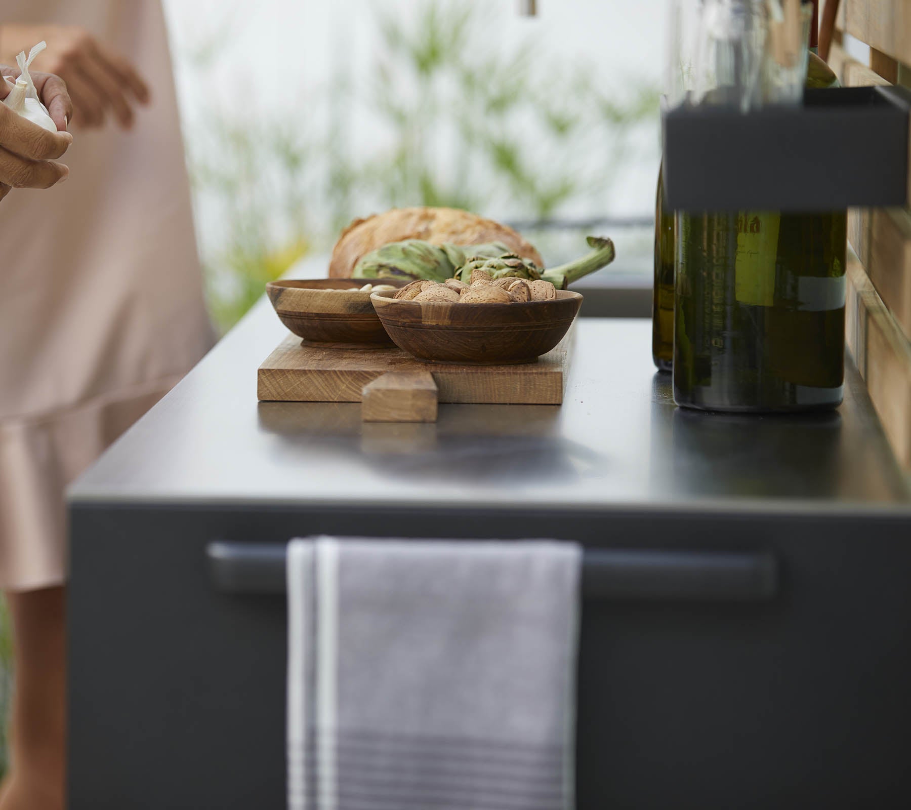 Un primer plano de una encimera de cocina con cuencos de madera de comida, una toalla y botellas de aceite de oliva, con vegetación suave al fondo.
