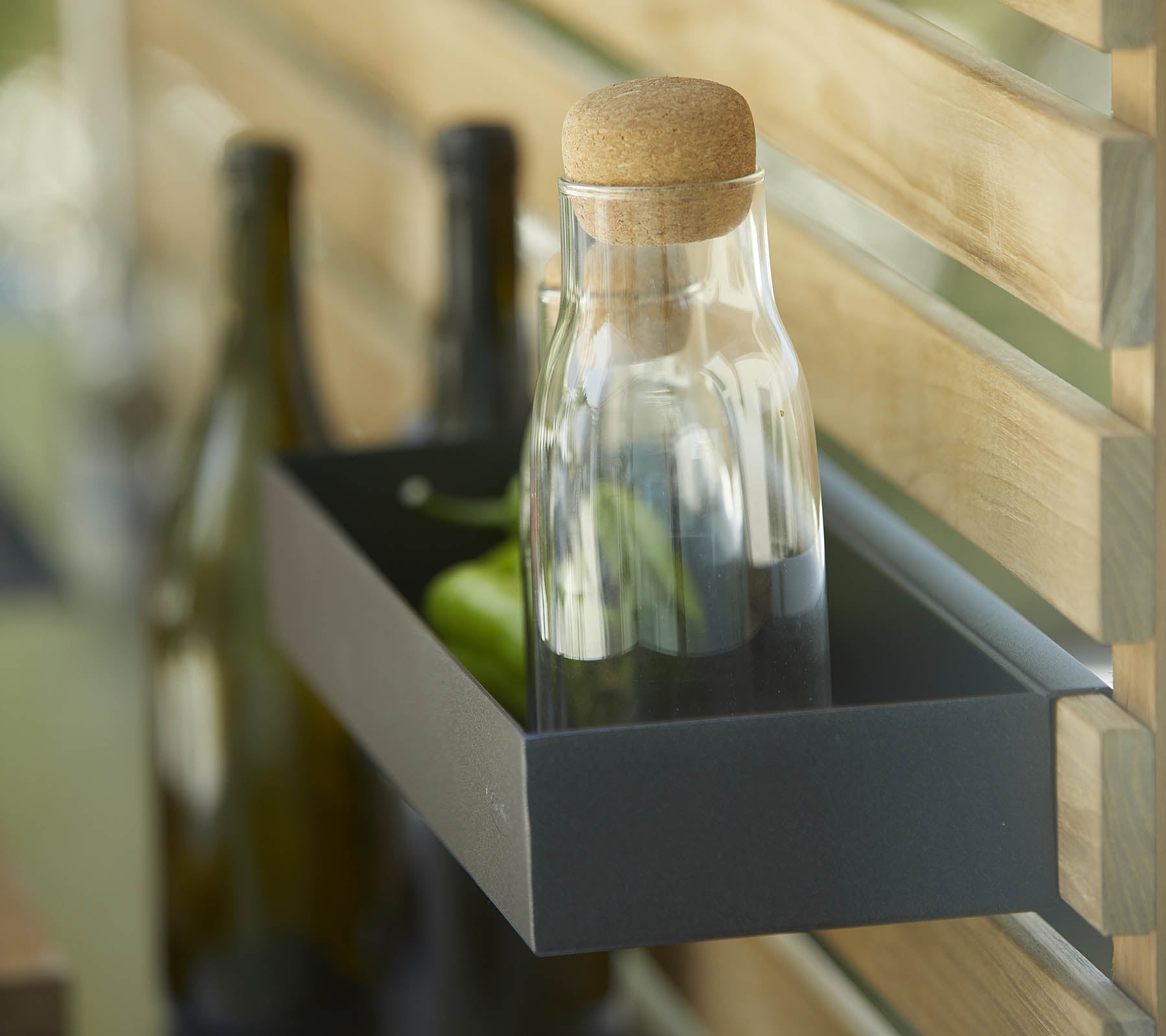 Una botella de vidrio con un tapón de corcho está en una estantería junto a varias botellas oscuras, contra un fondo de madera.