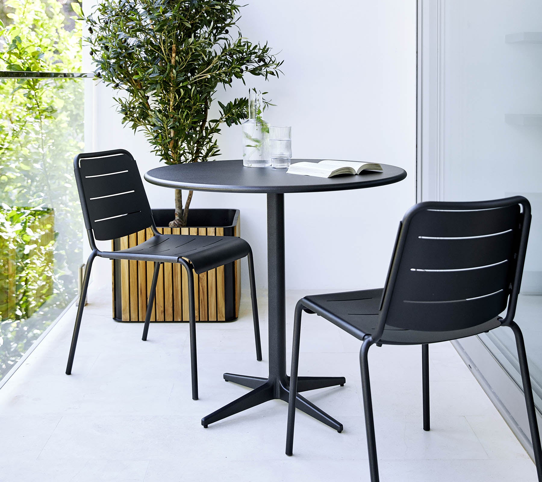 Pequeña mesa redonda con dos sillas negras y una planta en maceta cerca, creando un ambiente minimalista y elegante.