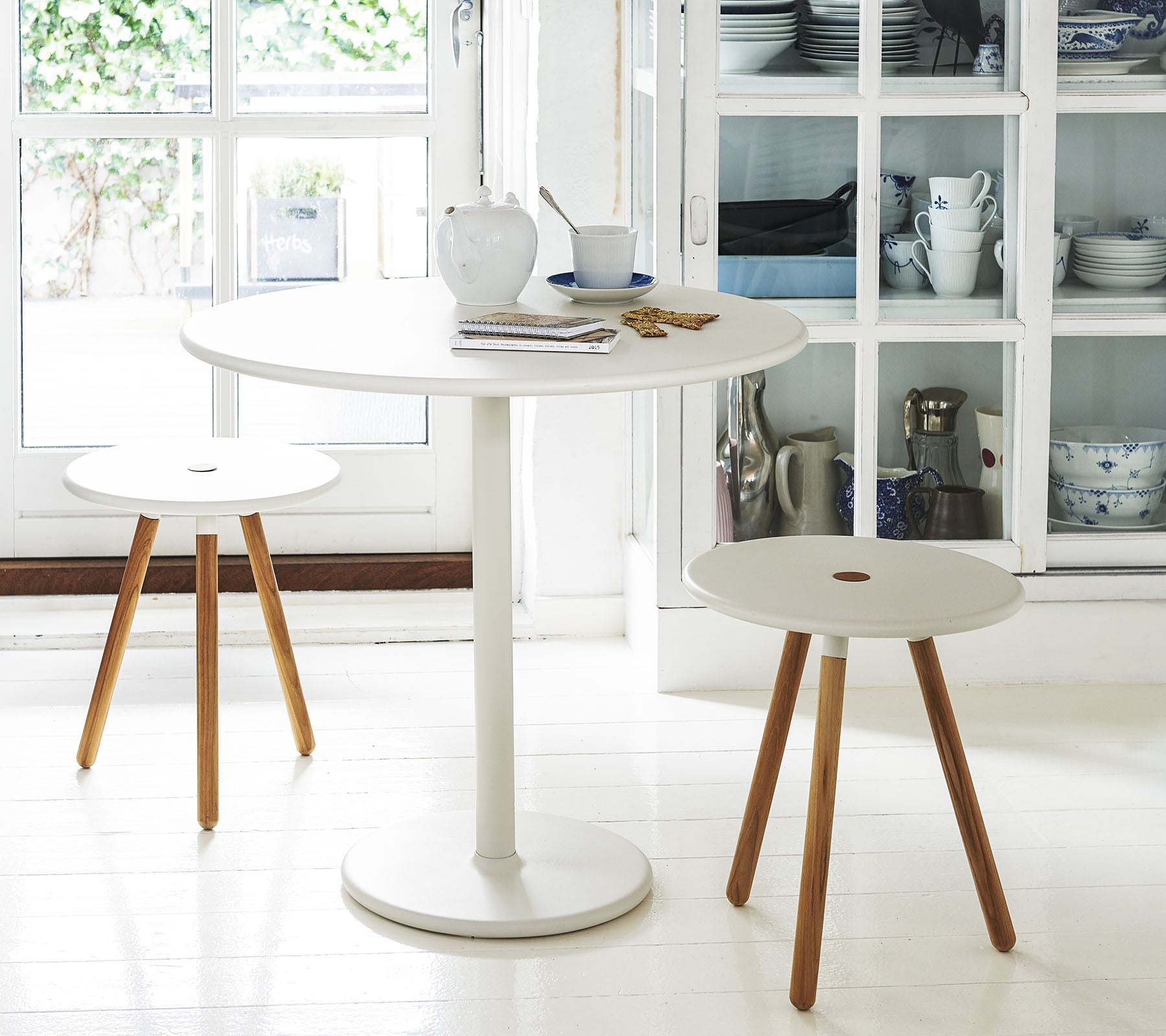 Una mesa blanca redonda acompañada de dos taburetes minimalistas con patas de madera, situada frente a un fondo de estanterías con utensilios de cocina.