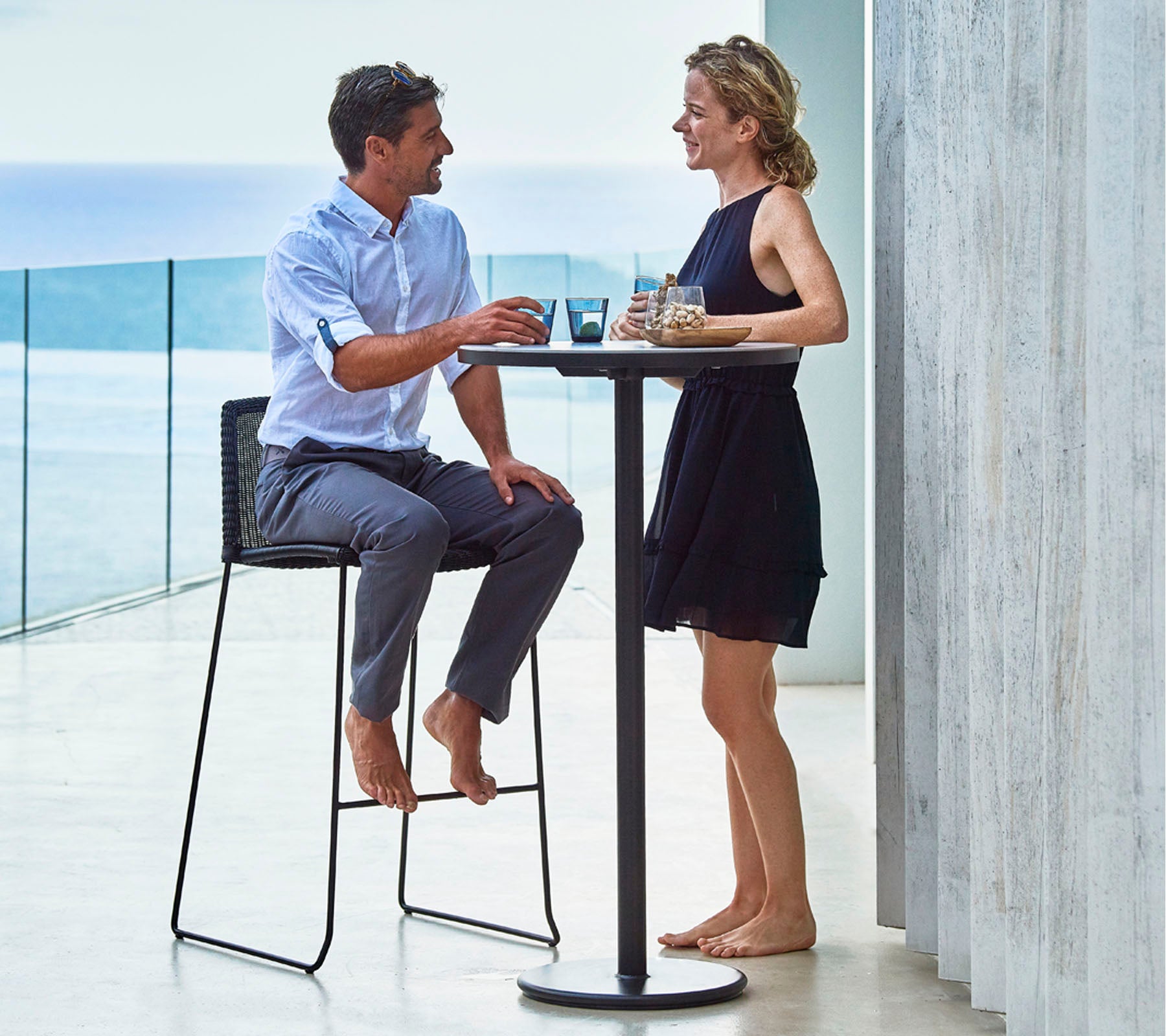 Un hombre y una mujer disfrutando de bebidas en una mesa alta, involucrados en una conversación, con un paisaje pintoresco de agua a lo lejos.