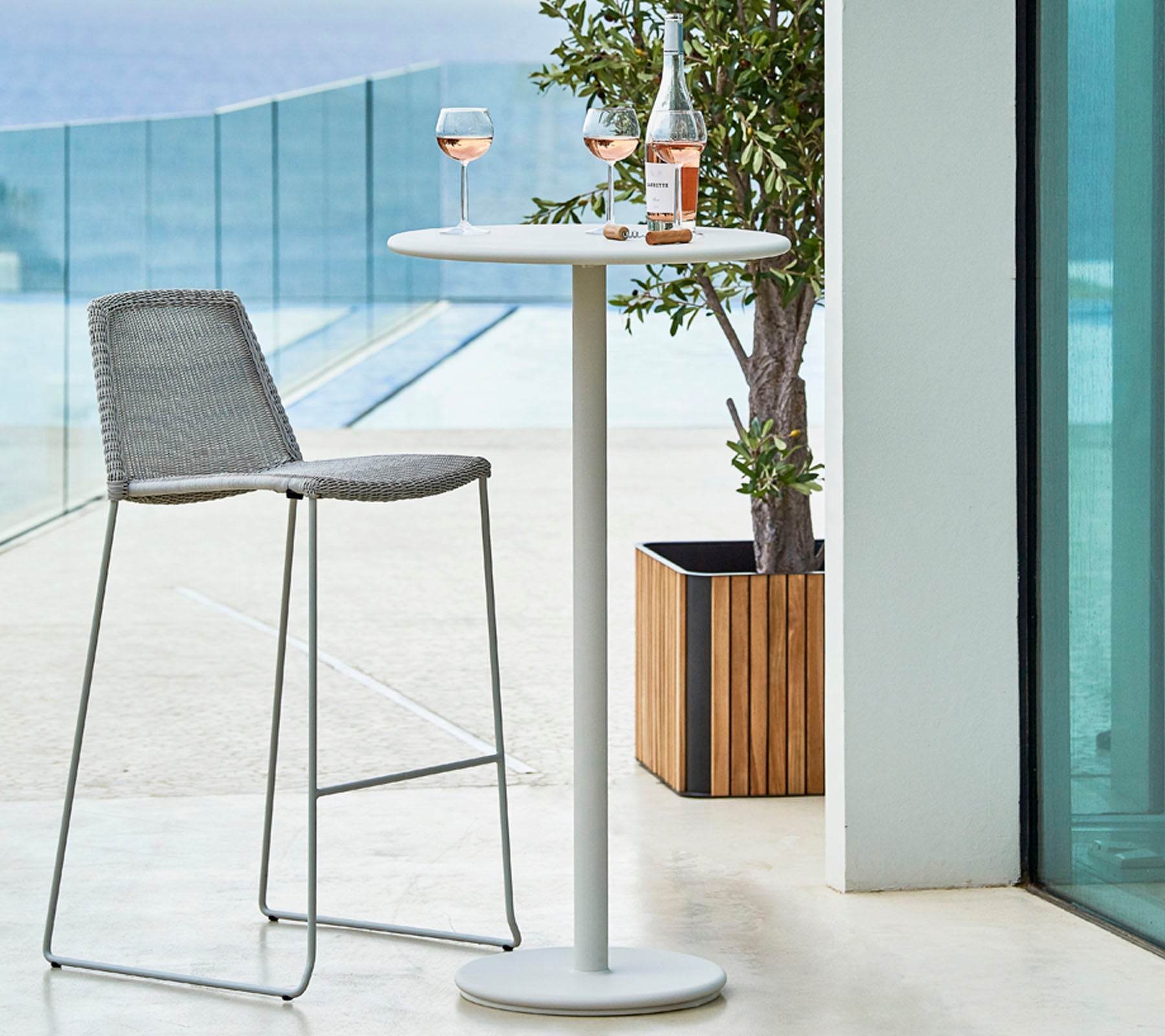Configuración exterior elegante con una mesa alta, un taburete de bar gris y dos copas de rosado acompañadas de una botella en la mesa.