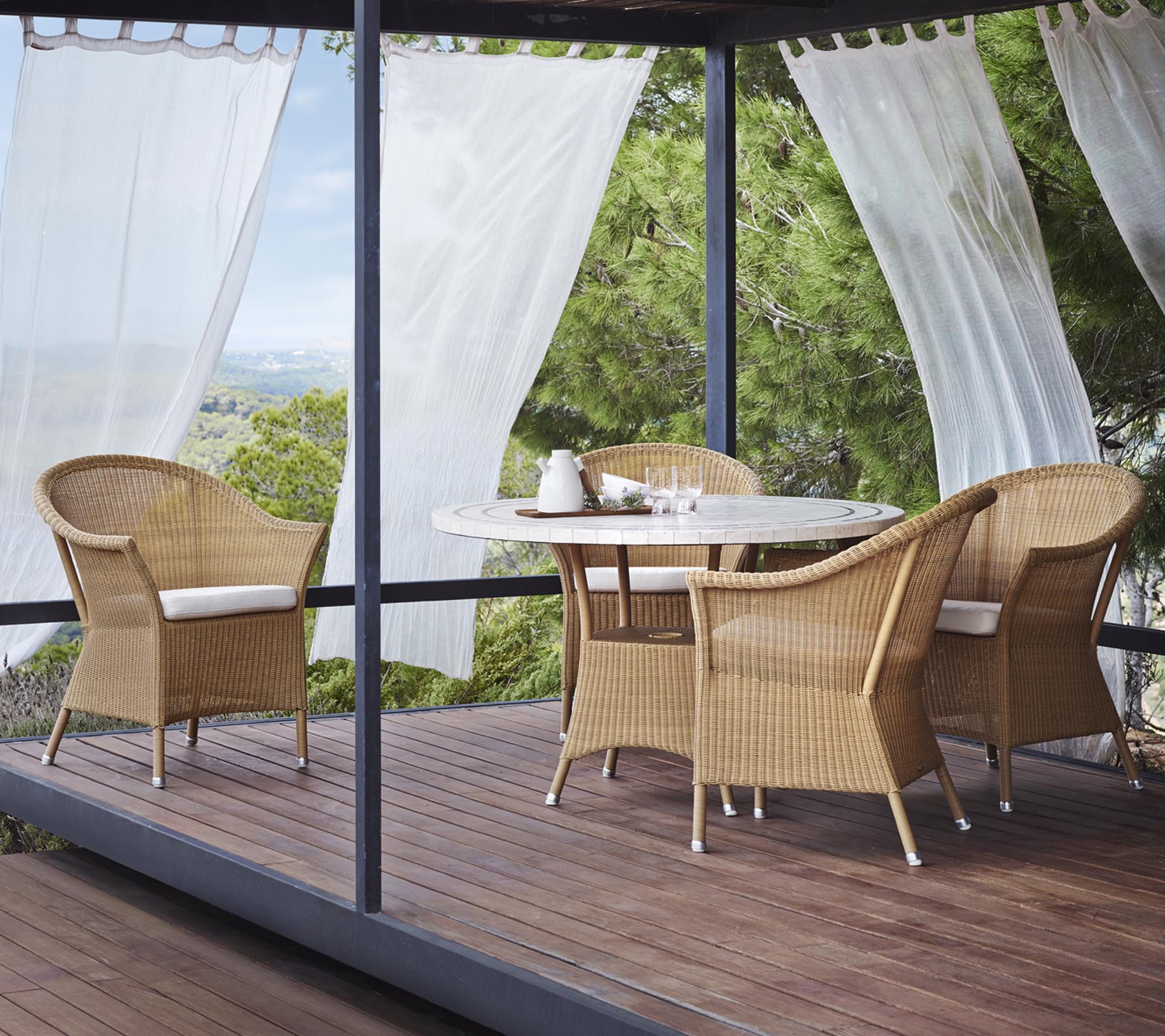 Una zona de estar elegante cuenta con cuatro sillas de mimbre y una mesa redonda, rodeada de cortinas ligeras y vegetación natural.