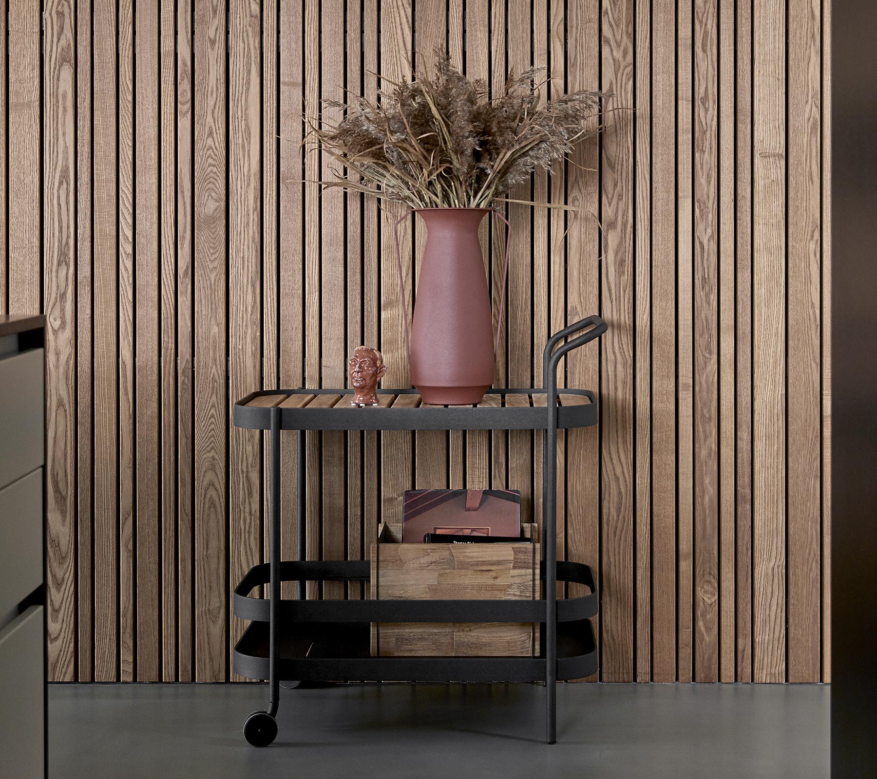 Un carro elegante con un acabado oscuro, que cuenta con cajas de almacenamiento de madera y un jarrón rosa con flores secas.