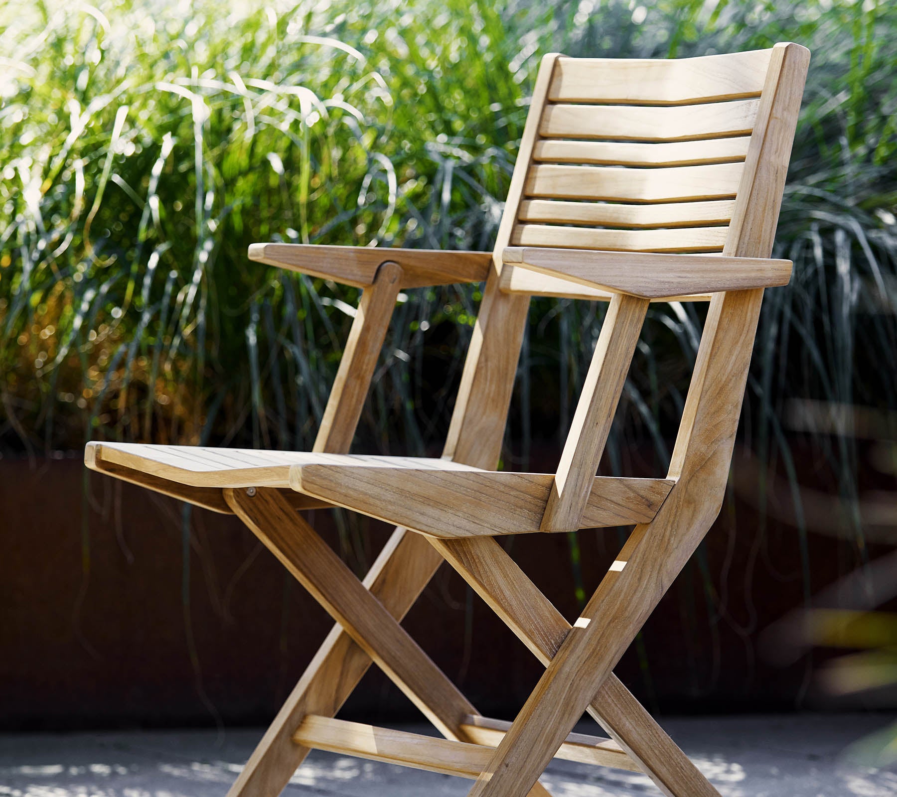 Una silla plegable de madera con detalles de listones y reposabrazos, rodeada de frondosa vegetación verde. Ideal para la relajación y entornos al aire libre.