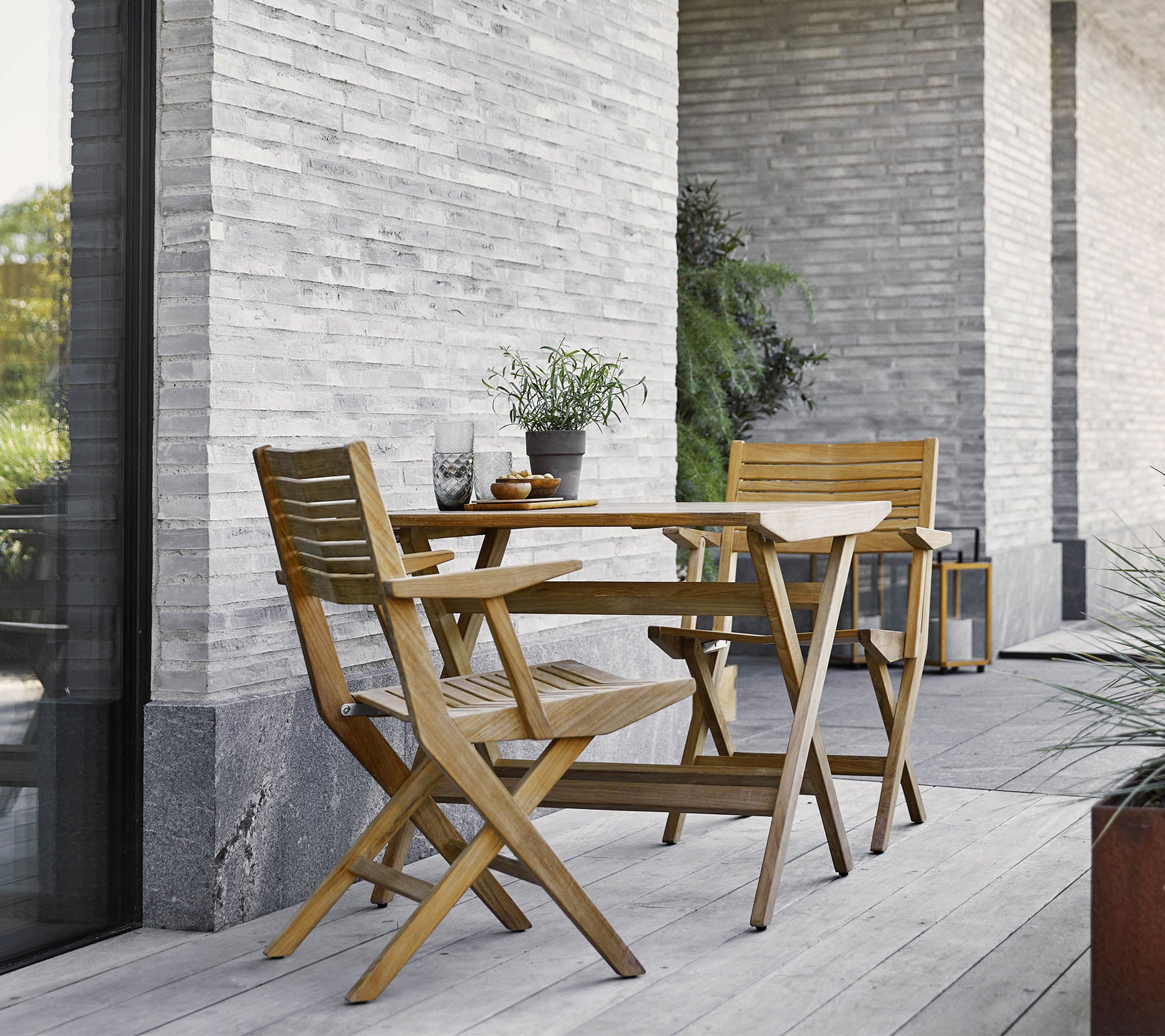 Una mesa de madera plegable con dos sillas a juego, adornada con plantas en macetas, contra una pared de piedra texturizada.