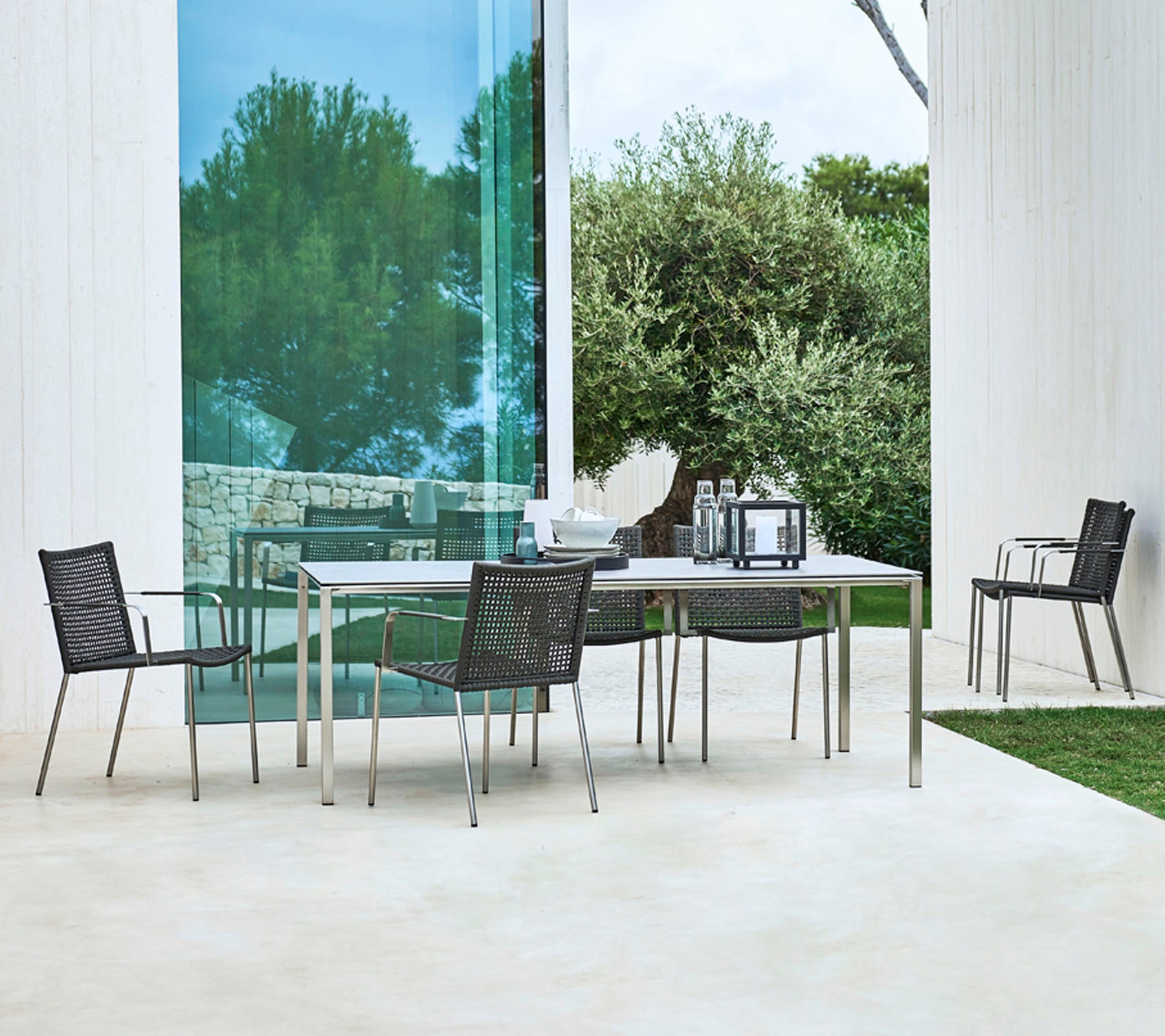 Un conjunto de comedor al aire libre moderno que cuenta con una mesa rectangular y cuatro sillas elegantes con un diseño aerodinámico, rodeado de vegetación.