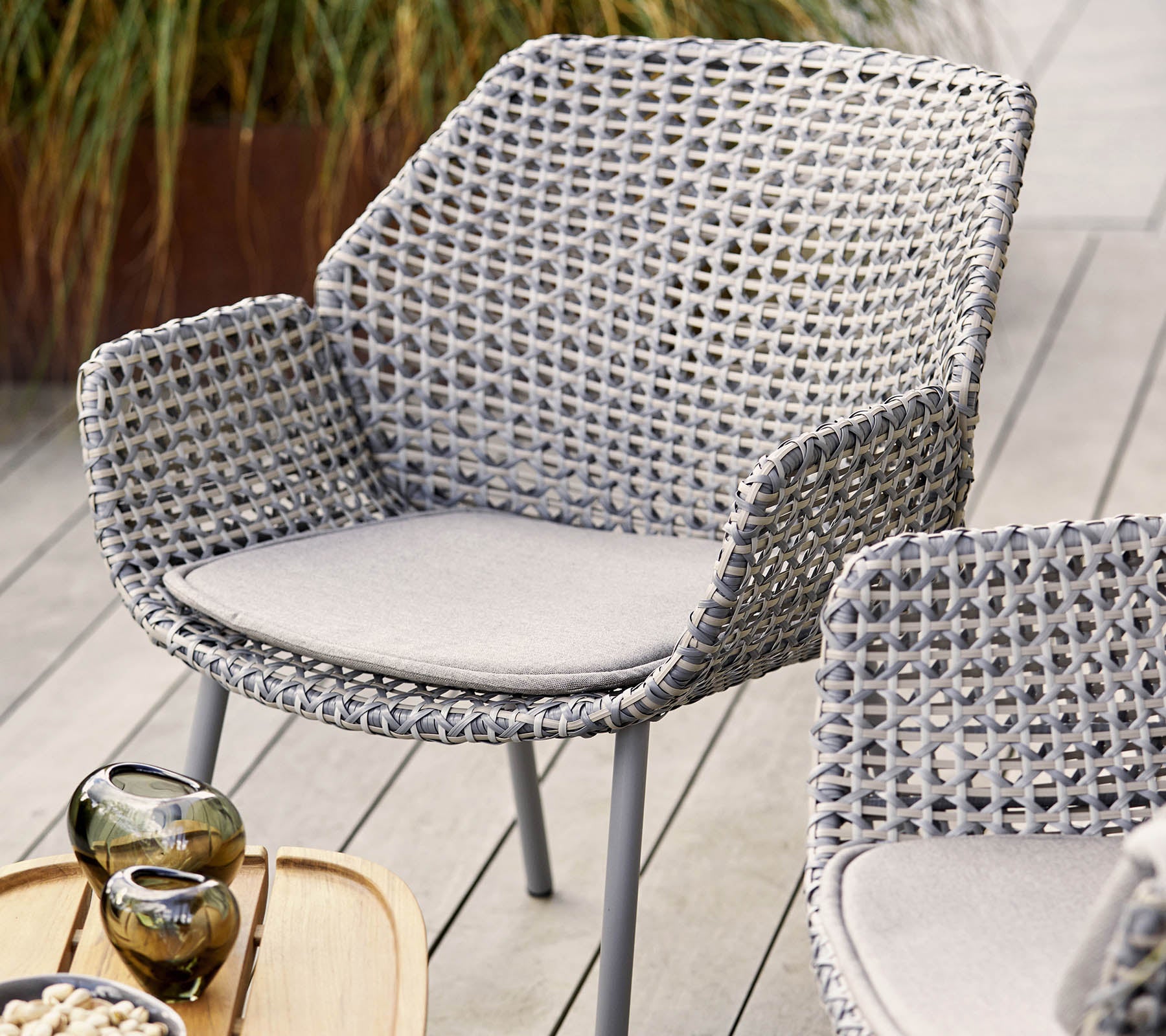 Silla tejida elegante en gris claro con un cojín cómodo, complementada con jarrones decorativos y una bandeja de madera.