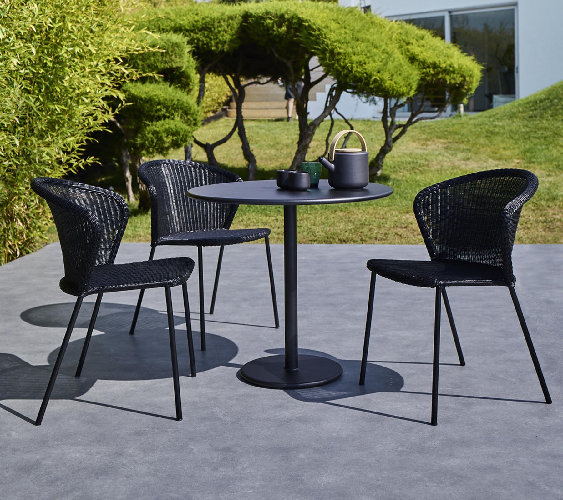 Un conjunto de mesa de exterior moderno que incluye una mesa redonda y tres sillas trenzadas negras, completo con una tetera y tazas sobre la mesa.