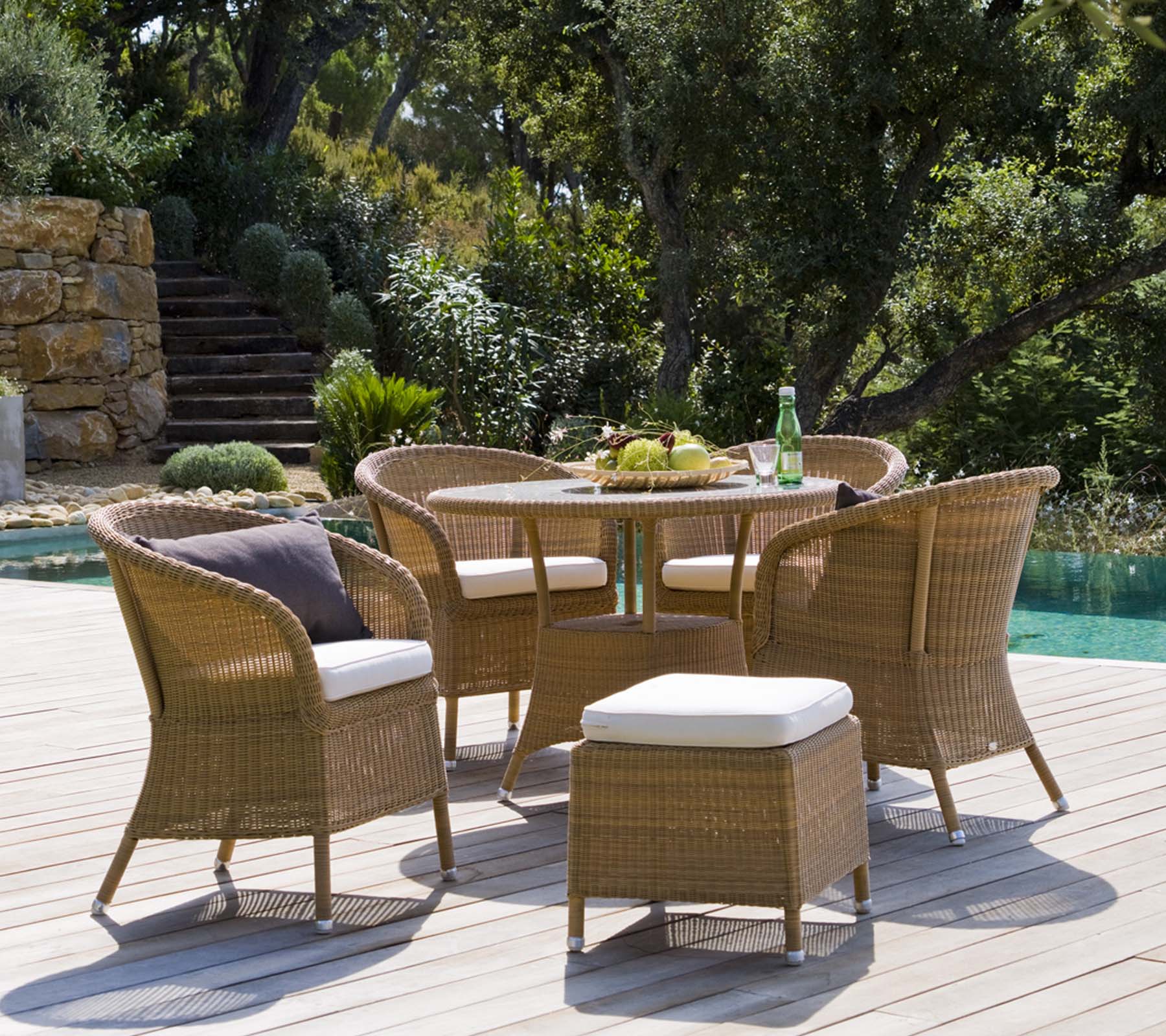 Un conjunto de asientos al aire libre elegante con una mesa redonda, cuatro sillas trenzadas y un pouf, rodeado de exuberante vegetación y un ambiente tranquilo.