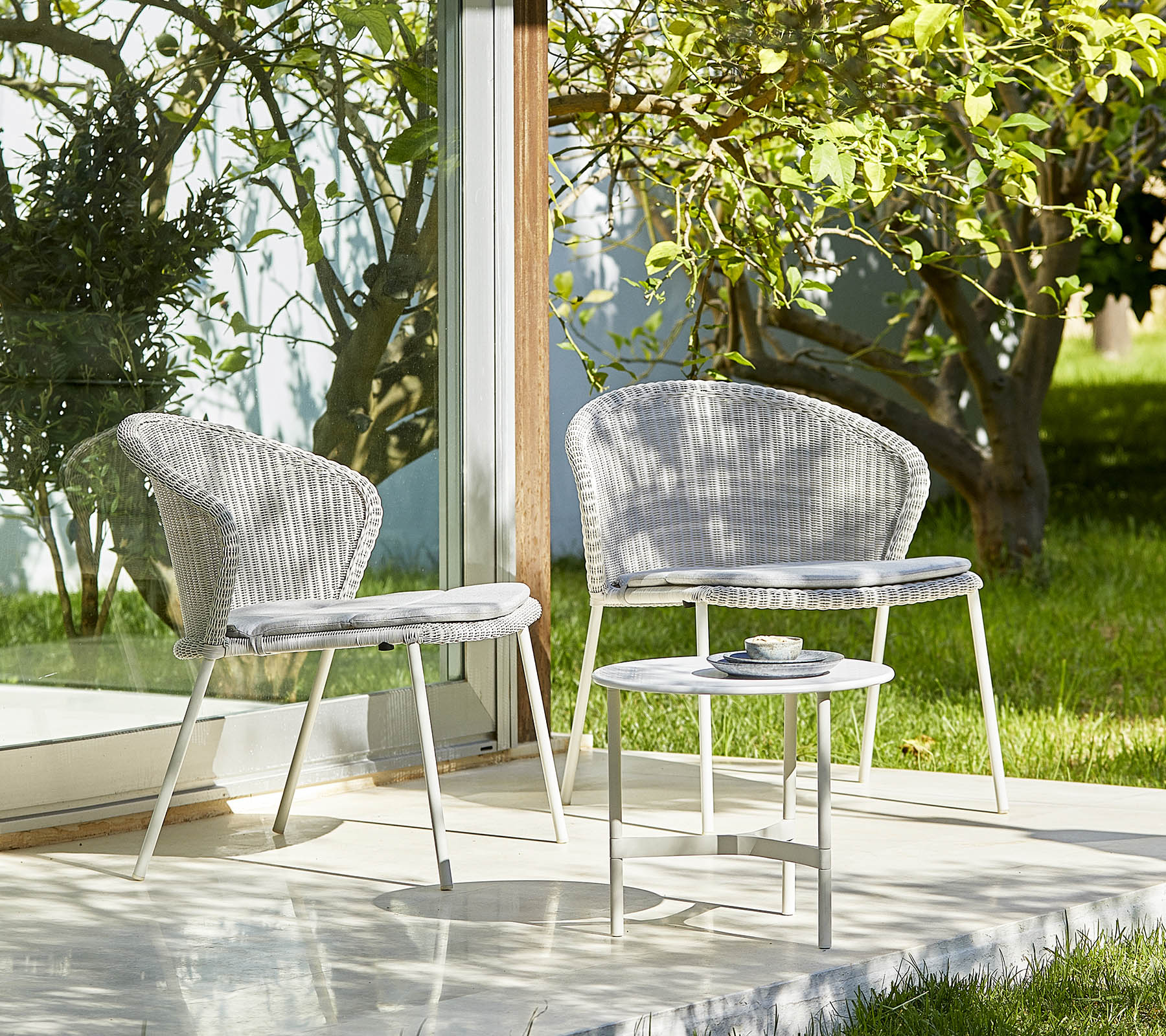 Dos sillas elegantes y una mesa pequeña en un entorno sereno con vegetación, perfecto para relajarse y reuniones al aire libre.