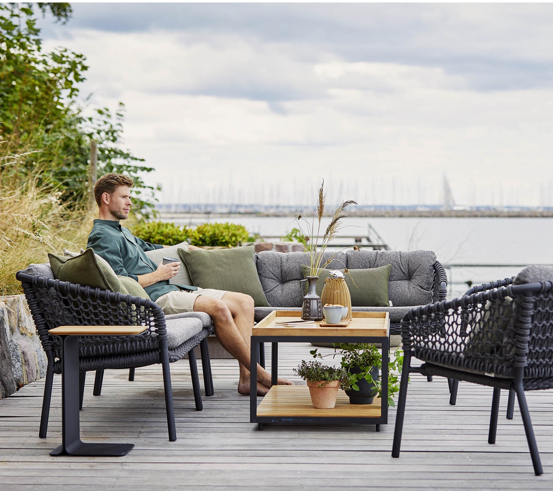 Una zona de estar cómoda con un hombre relajándose, con sillas de mimbre elegantes y una mesa de café de madera adornada con plantas y objetos decorativos.