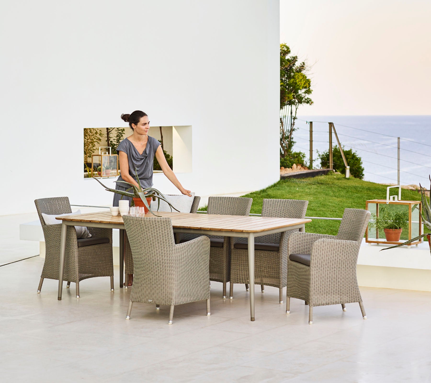 Una mujer prepara la mesa del comedor rodeada de sillas de mimbre, con vista a un paisaje pintoresco con vegetación y agua de fondo.