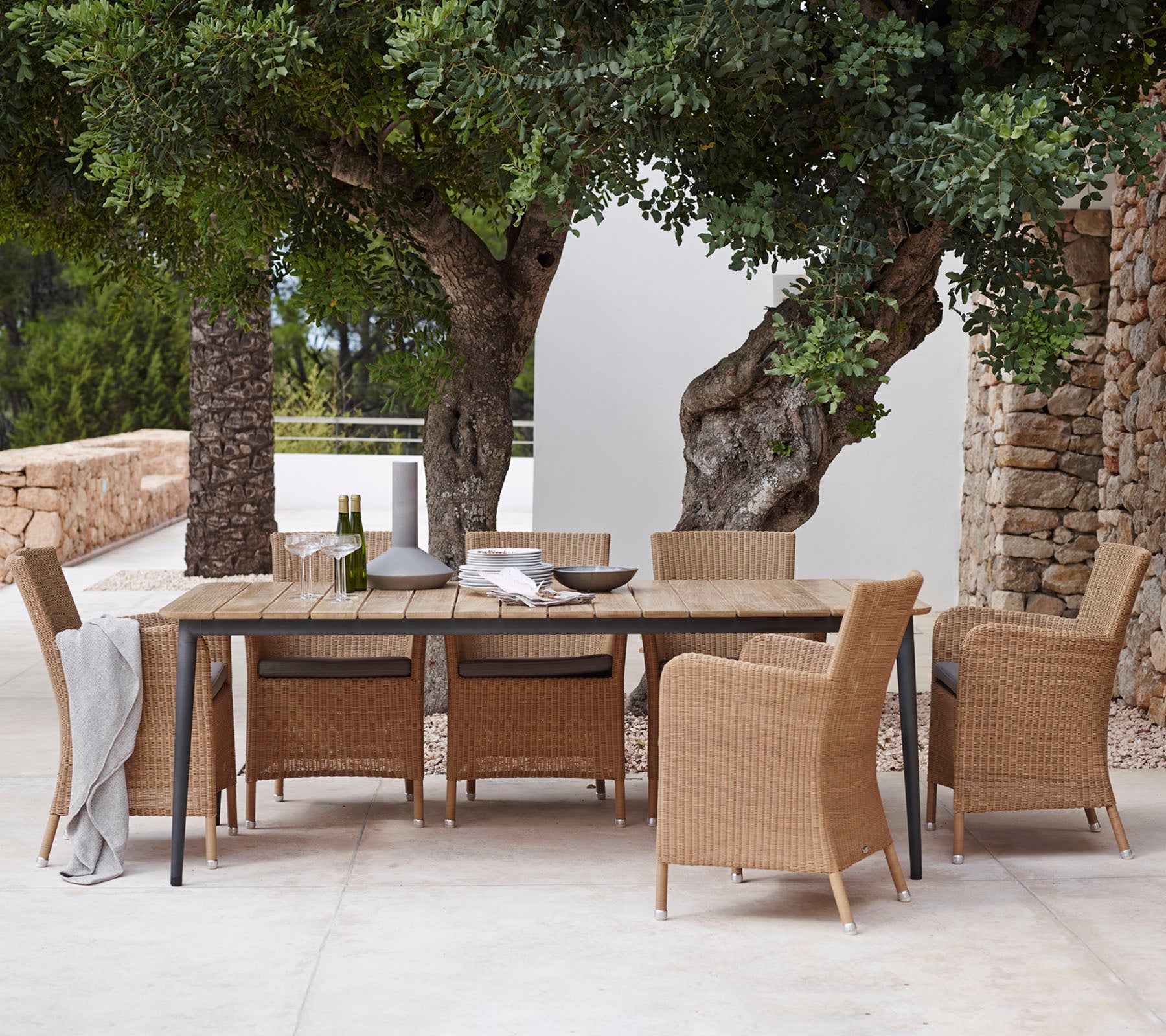 Un conjunto de comedor elegante con una mesa larga y sillas tejidas, situado bajo una vegetación exuberante y rodeado de paredes de piedra natural.