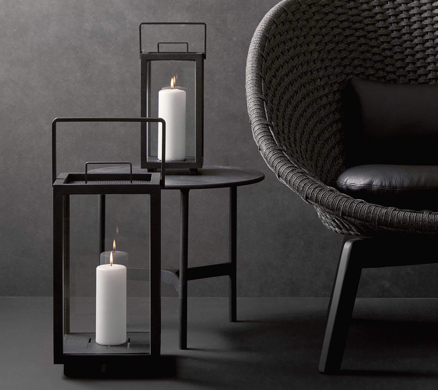 Dos faroles elegantes con velas blancas, acompañados de una silla moderna y texturizada sobre una mesa minimalista contra un fondo oscuro.