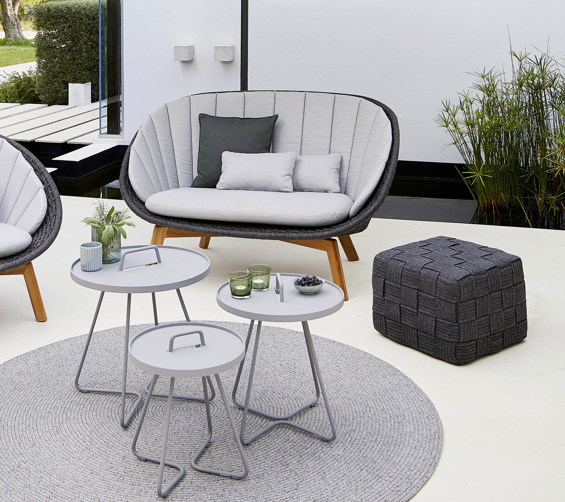 Área de descanso acogedora con un sofá elegante, un par de mesas redondas, un pouf decorativo y una suave alfombra, rodeada de vegetación.