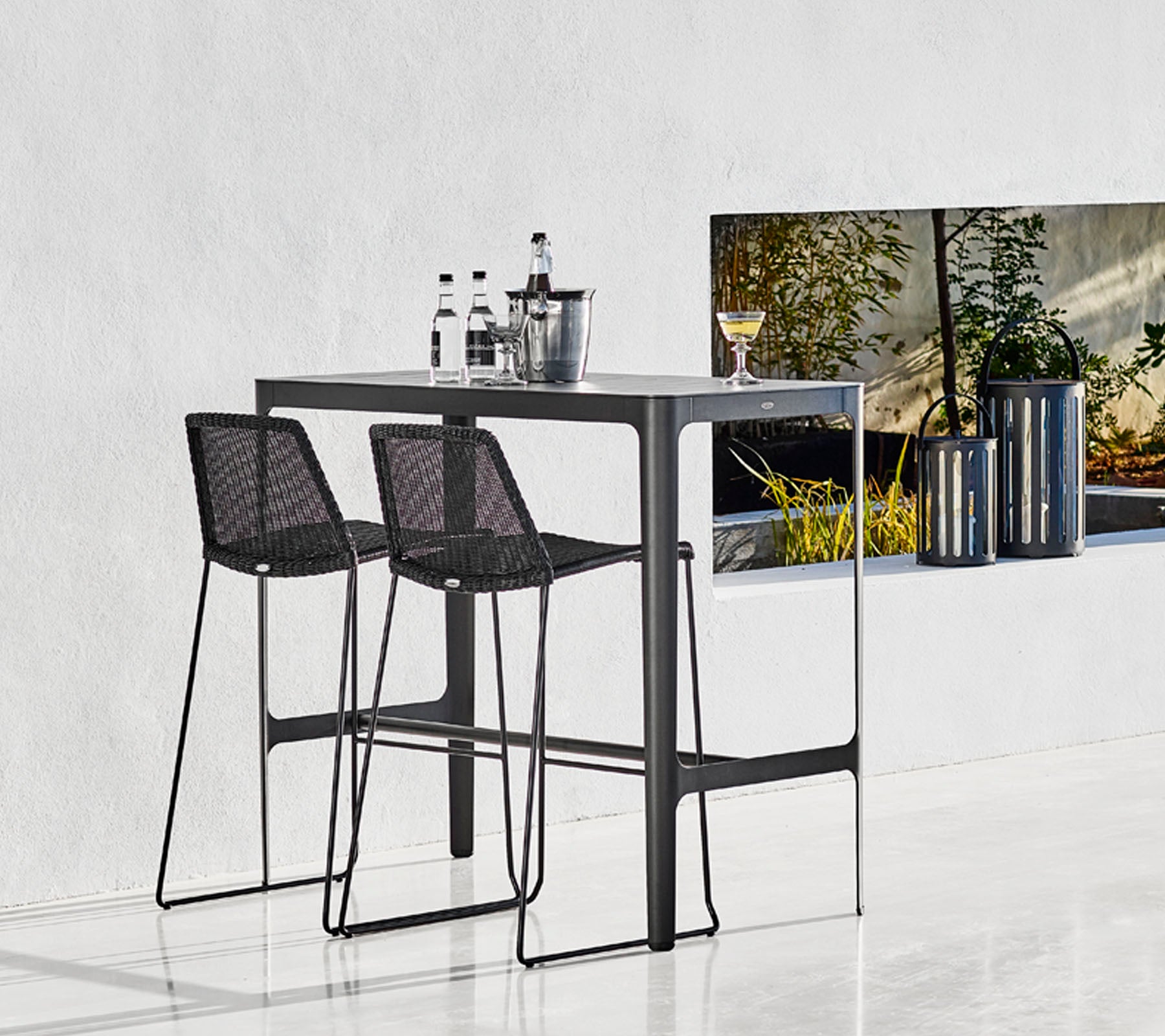 Mesa de bar elegante con dos taburetes de mimbre negros, acompañada de cristalería y un cubo elegante, con un fondo minimalista.