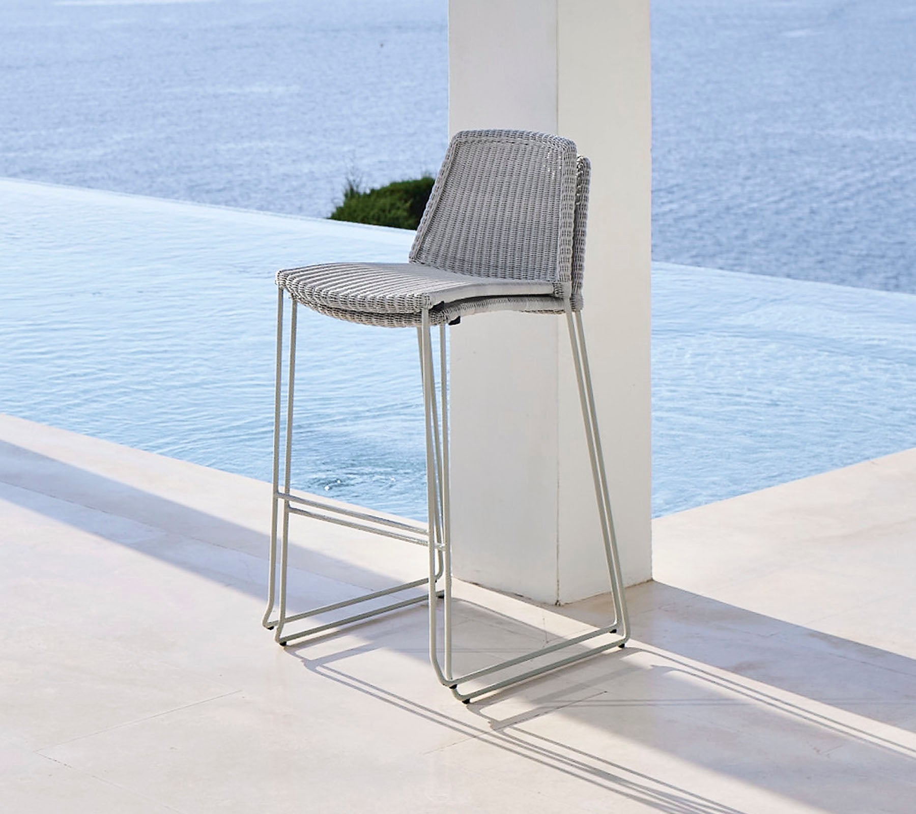 Taburete de bar elegante con un asiento de mimbre y patas metálicas elegantes, situado cerca de una característica de agua tranquila.