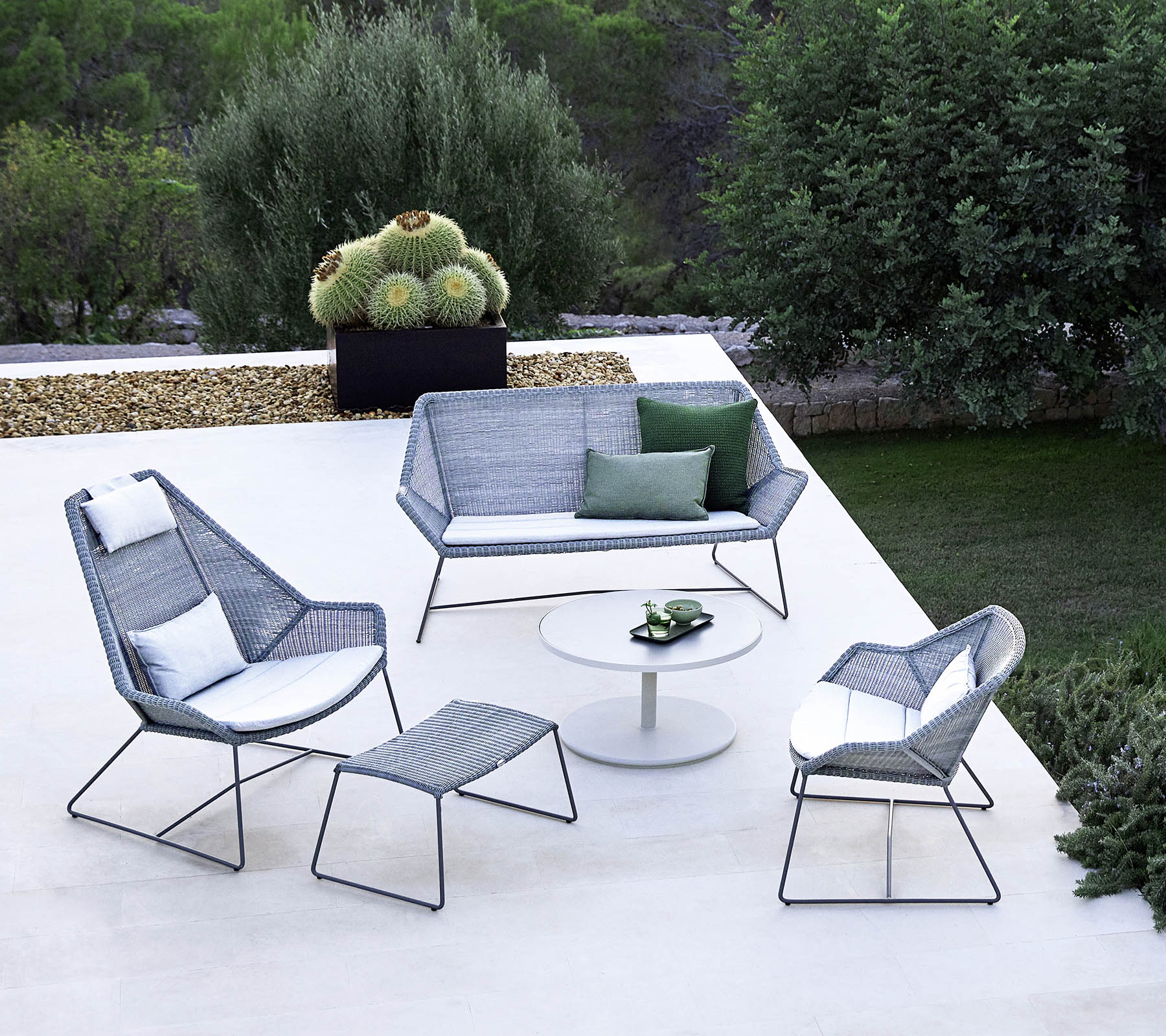 Elegante disposición de asientos al aire libre que incluye un sofá, dos sillones, un reposapiés y una mesa de café, complementada con vegetación en el fondo.
