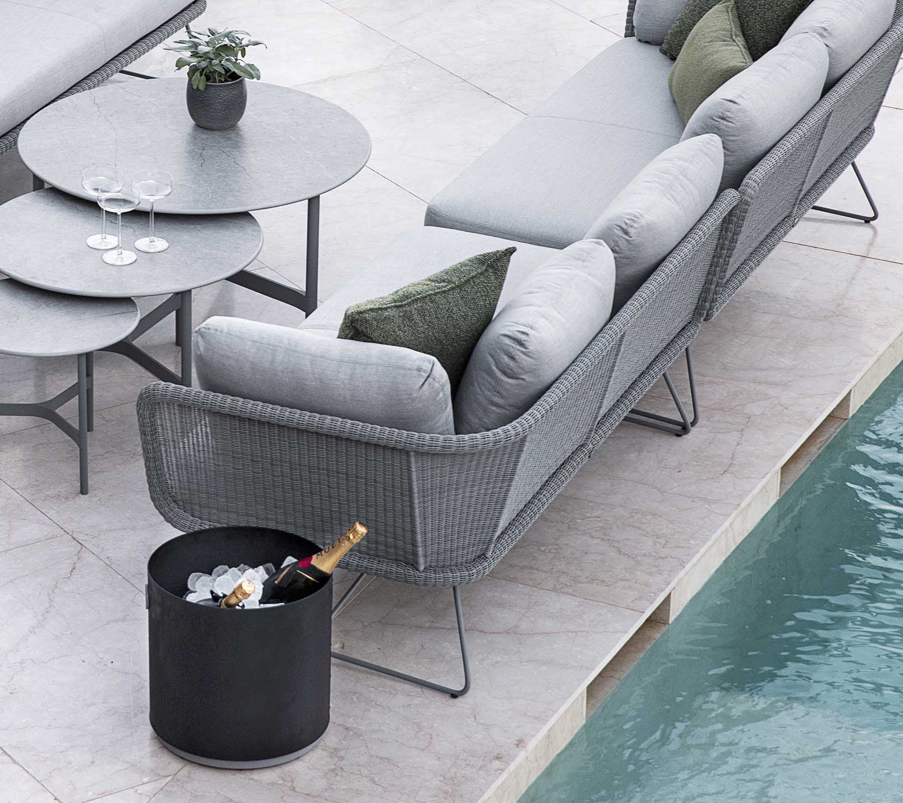 Conjunto moderno de asientos al aire libre con un elegante sofá gris y una mesa redonda, complementado con un cubo de hielo y bebidas.