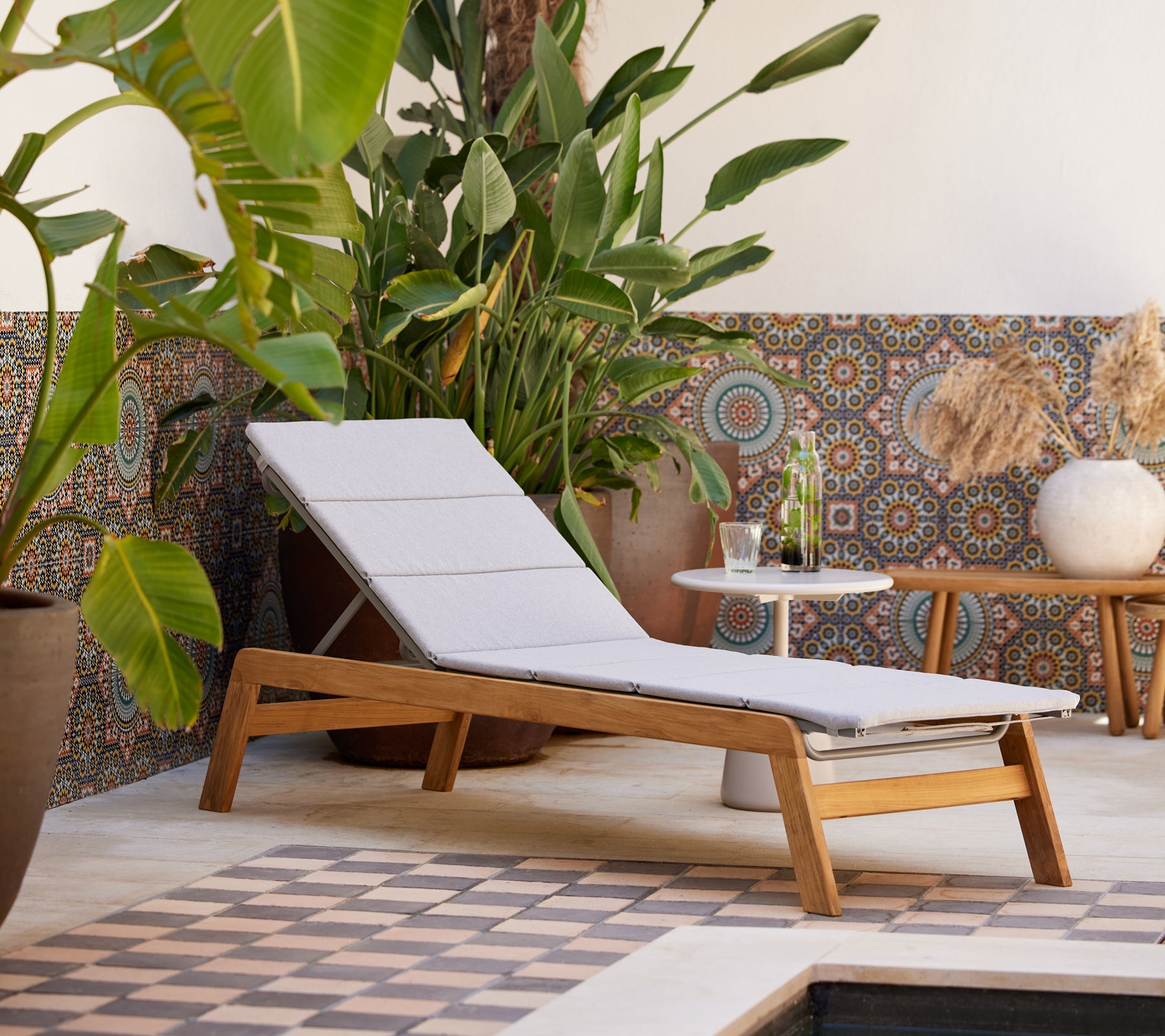 Una silla de lounge moderna con un cojín ligero, acompañada de una mesa auxiliar, rodeada de abundante vegetación y un fondo de azulejos decorativos.