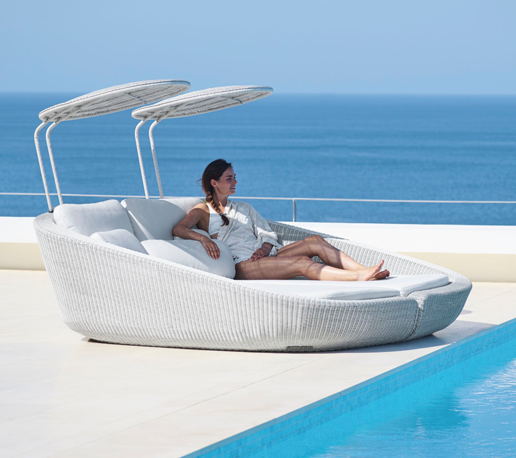 Una mujer se relaja en una silla de lounge moderna y estilosa con toldos ajustables junto a un cuerpo de agua tranquilo.