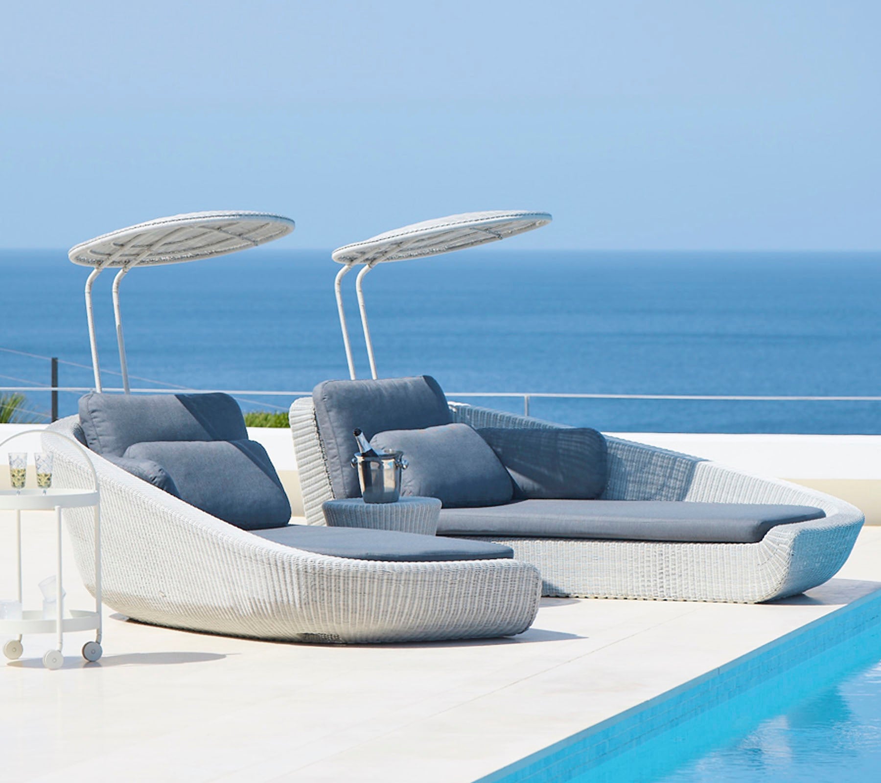 Sillas de lounge exteriores elegantes con toldos al lado de una piscina serena, con vistas a un mar tranquilo. Perfecto para relajarse y disfrutar del tiempo libre.