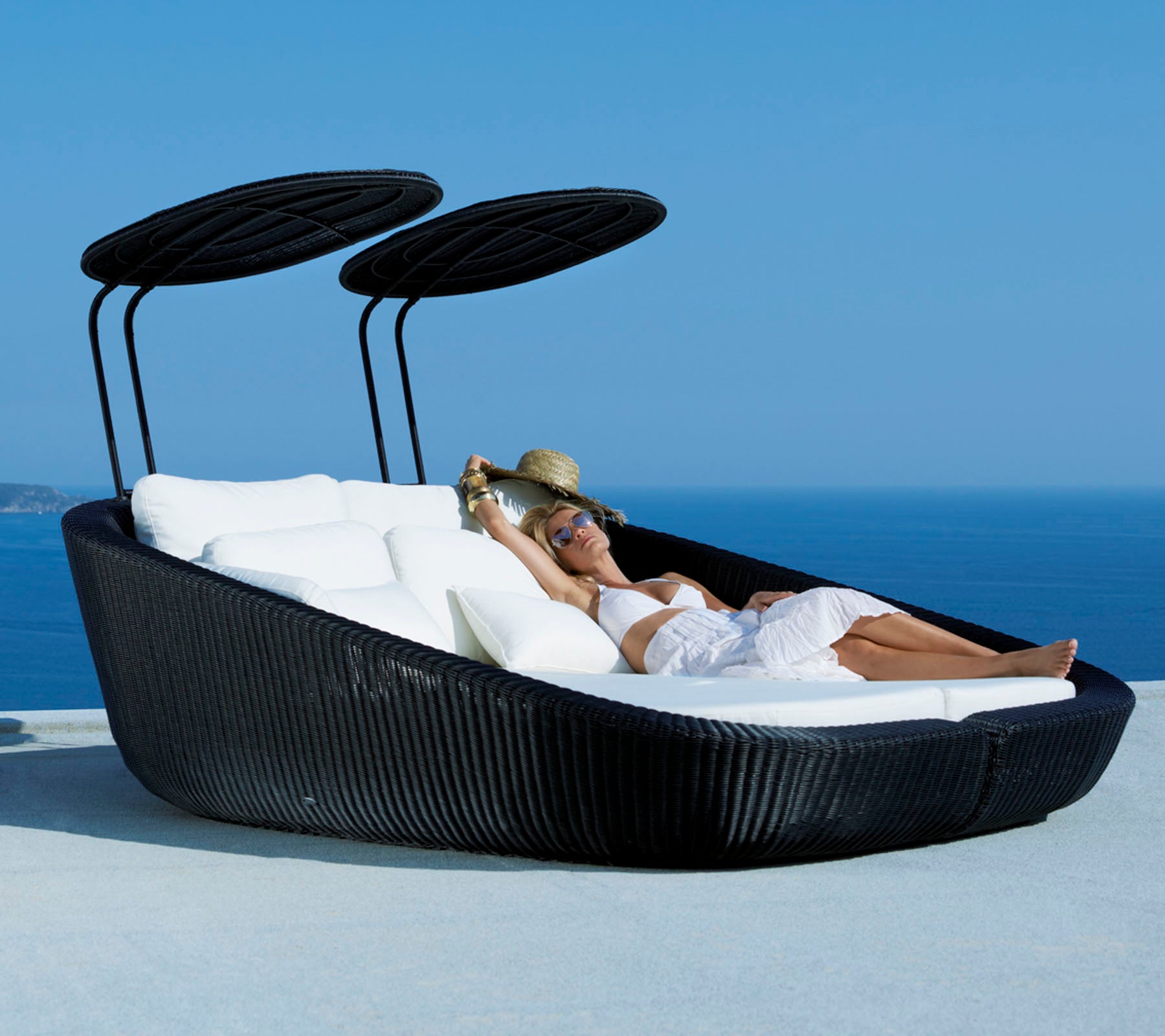 Una cama lounge exterior elegante con un diseño moderno, que cuenta con suaves cojines y toldos ajustables, con un fondo escénico del océano.