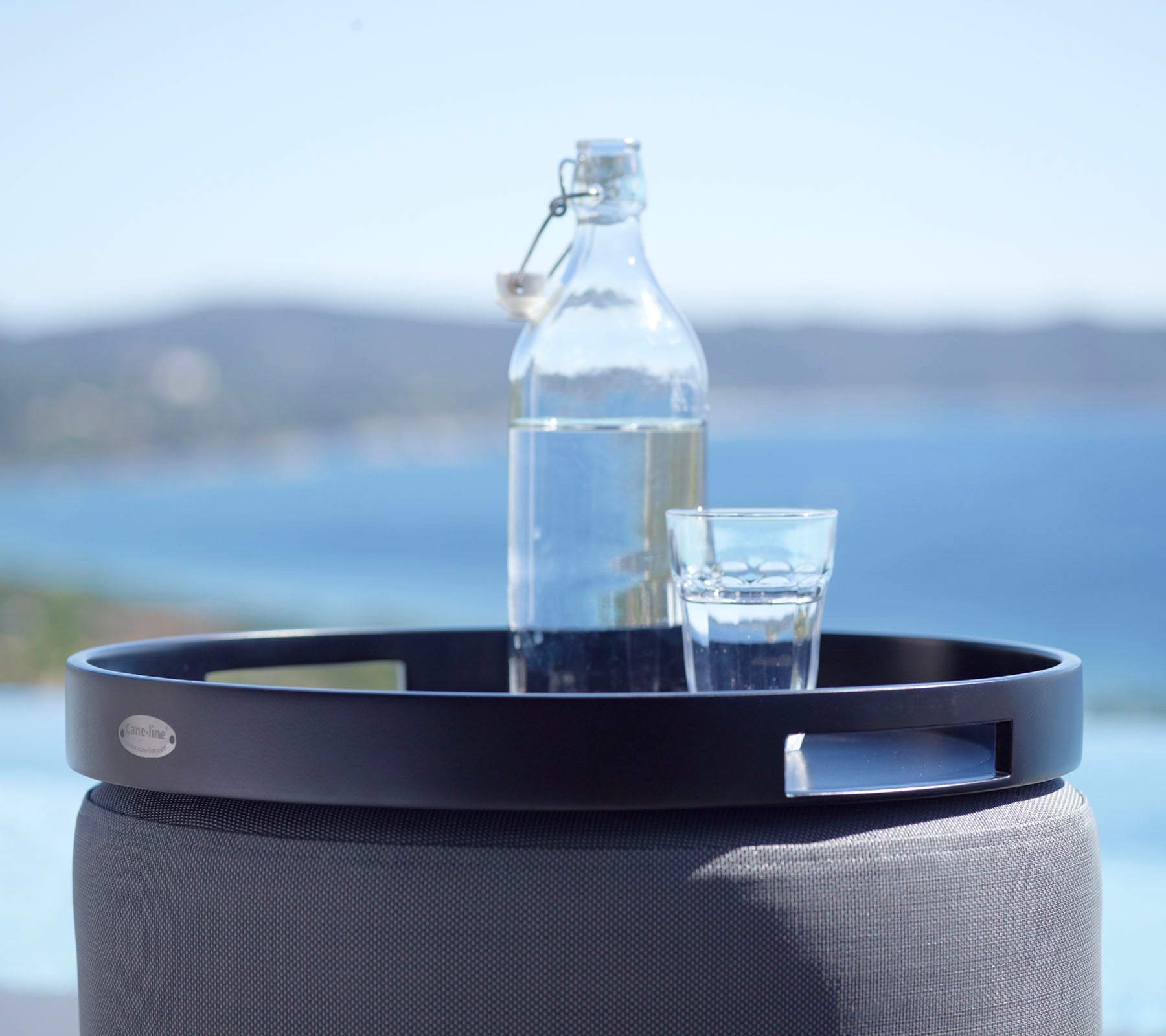 Una bandeja redonda negra sostiene un vaso de agua y una botella de vidrio, con un fondo escénico del mar y colinas distantes.