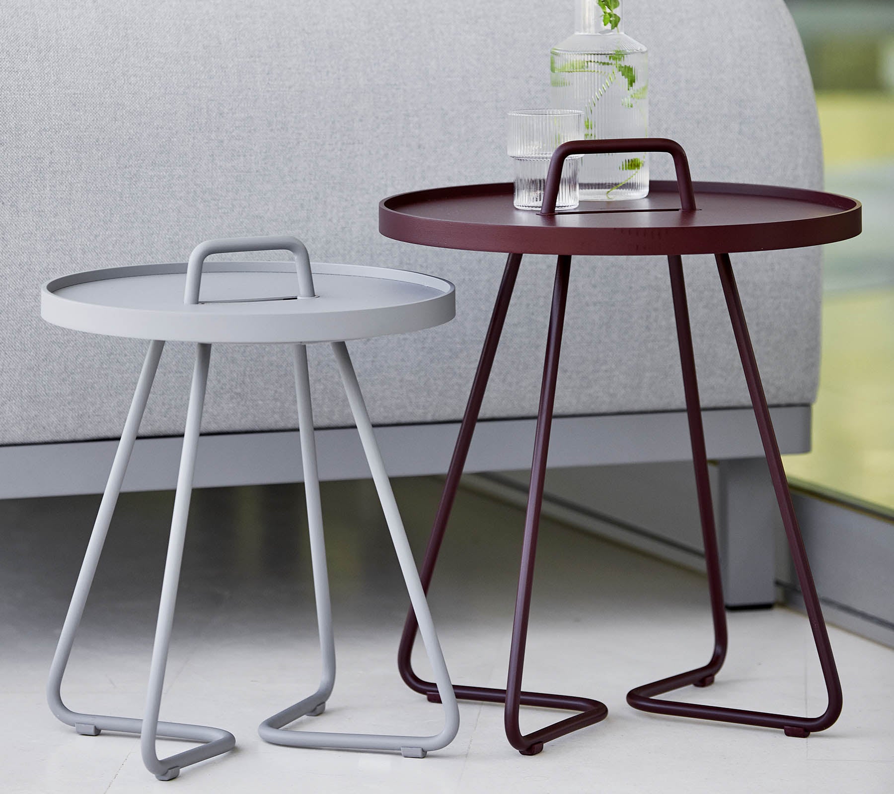 Dos mesas auxiliares modernas, una gris y una burdeos, ambas con bandejas redondas y elegantes patas de trípode. Ideal para decoración contemporánea.