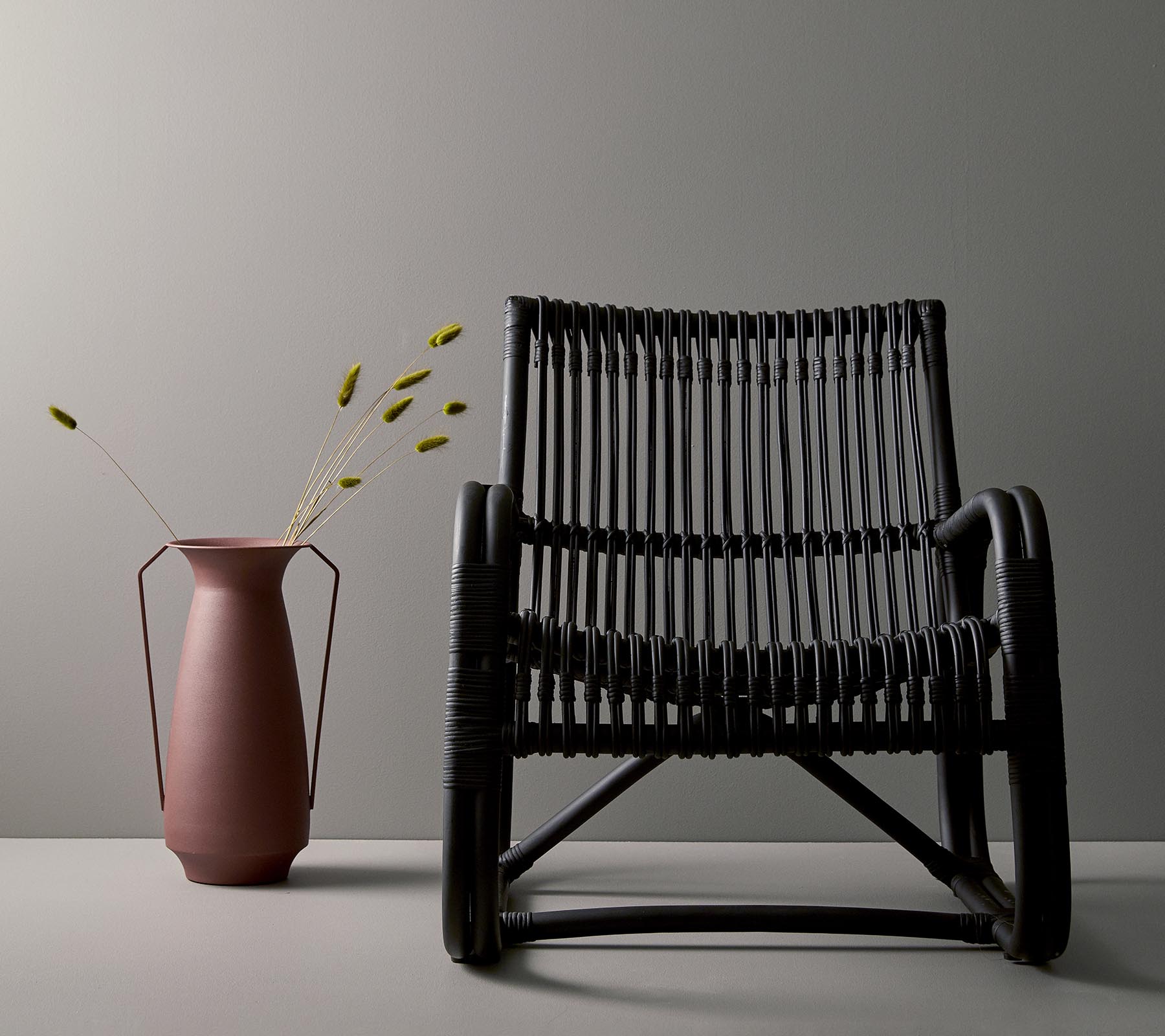 Silla negra tejida elegante al lado de un jarrón rosa con tallos verdes, contra un fondo gris minimalista.