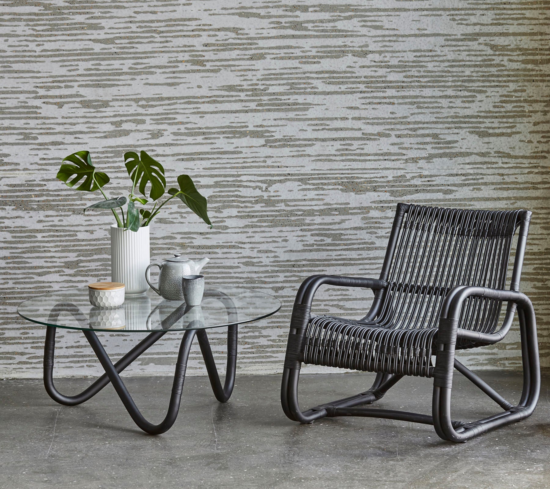 Una silla elegante al lado de una mesa de café de vidrio, con un jarrón con plantas y una pequeña taza de té, frente a una pared texturizada.