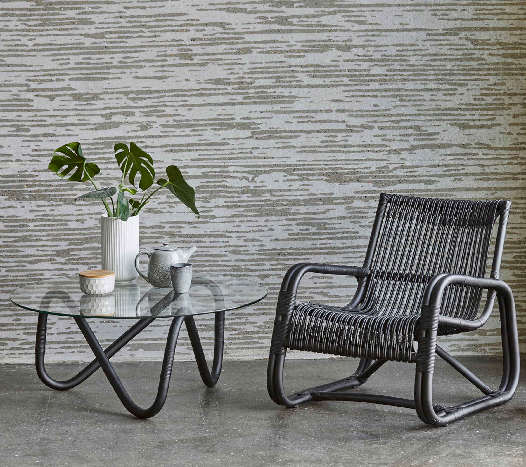 Una silla elegante y una mesa de cristal con una planta y un juego de café, complementada por un fondo de pared texturizada.
