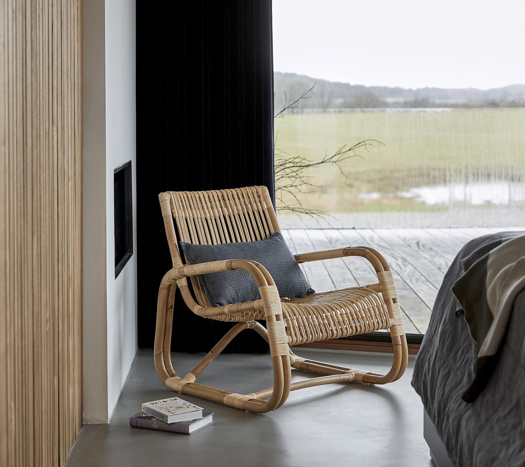 Una silla mecedora elegante hecha de materiales naturales, colocada al lado de una cama y un libro, con una vista escénica visible a través de grandes ventanas.