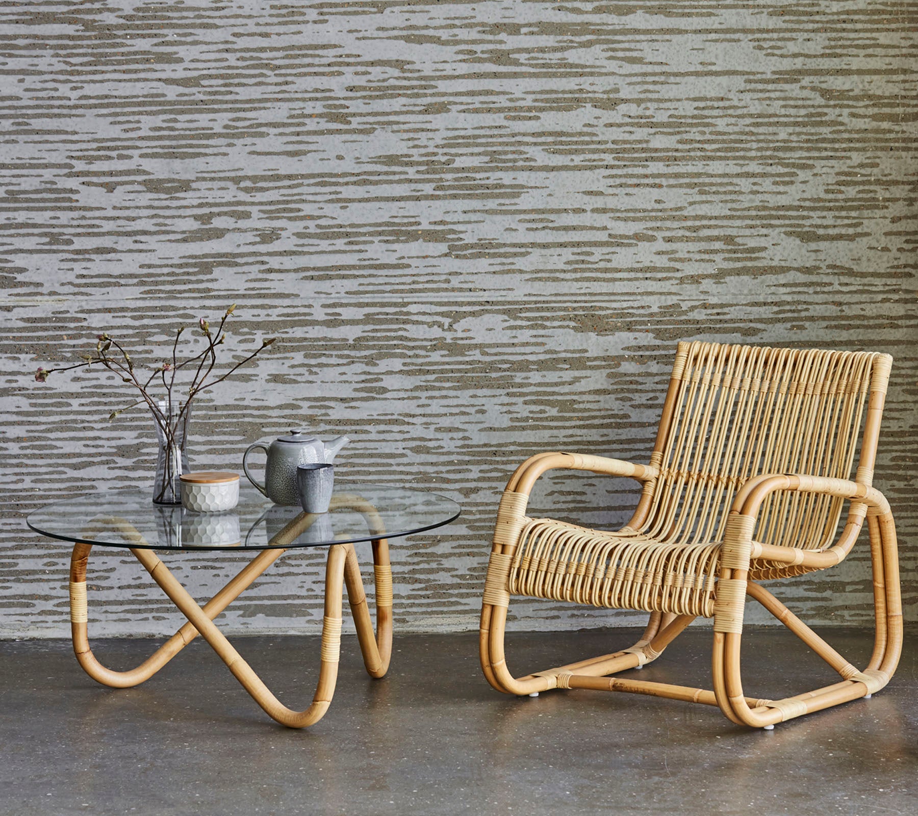 Una silla de ratán elegante junto a una mesa de café de vidrio, con un jarrón con ramitas y una pequeña tetera, situada contra un fondo texturizado.