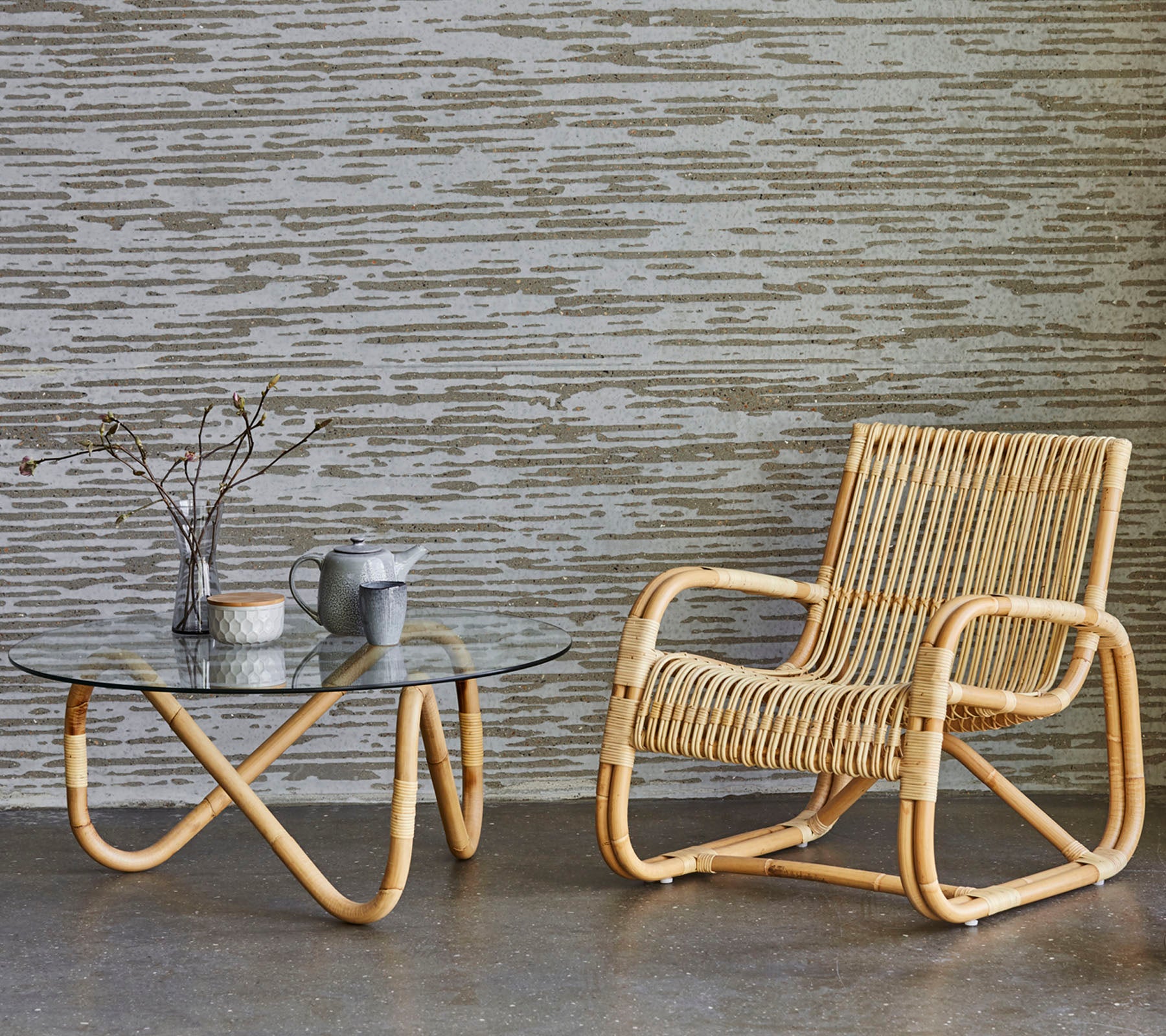 Silla de ratán y mesa de vidrio con objetos decorativos, situados contra una pared texturizada. Un estilo de decoración minimalista y moderno.