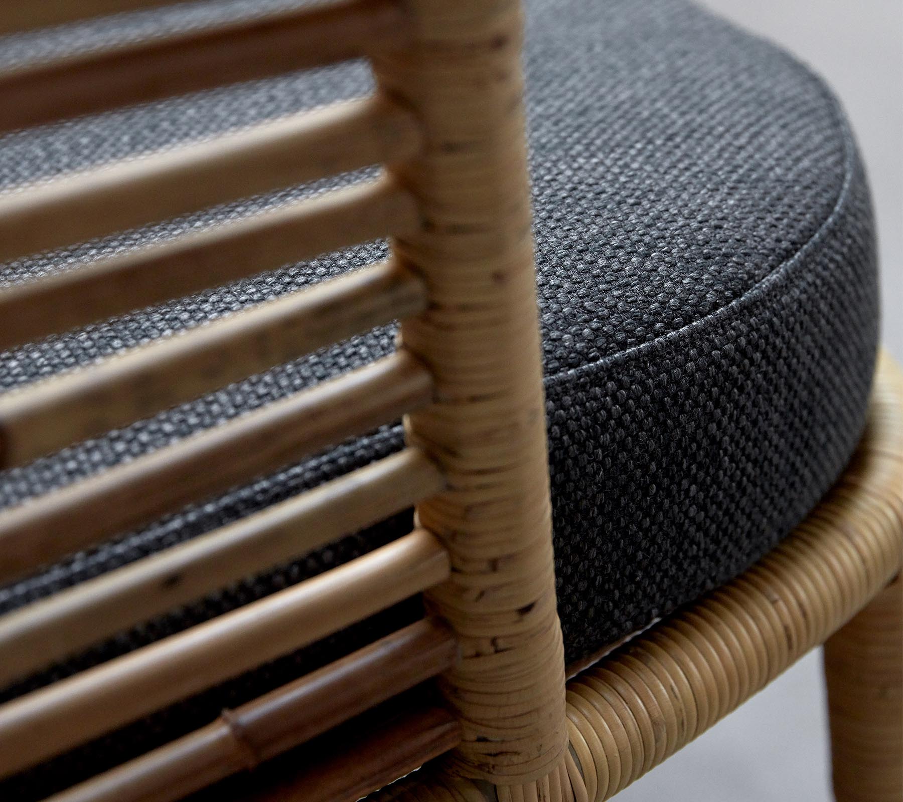 Primer plano de una silla tejida con un cojín redondo de color gris oscuro y un marco de madera natural que muestra detalles intrincados.