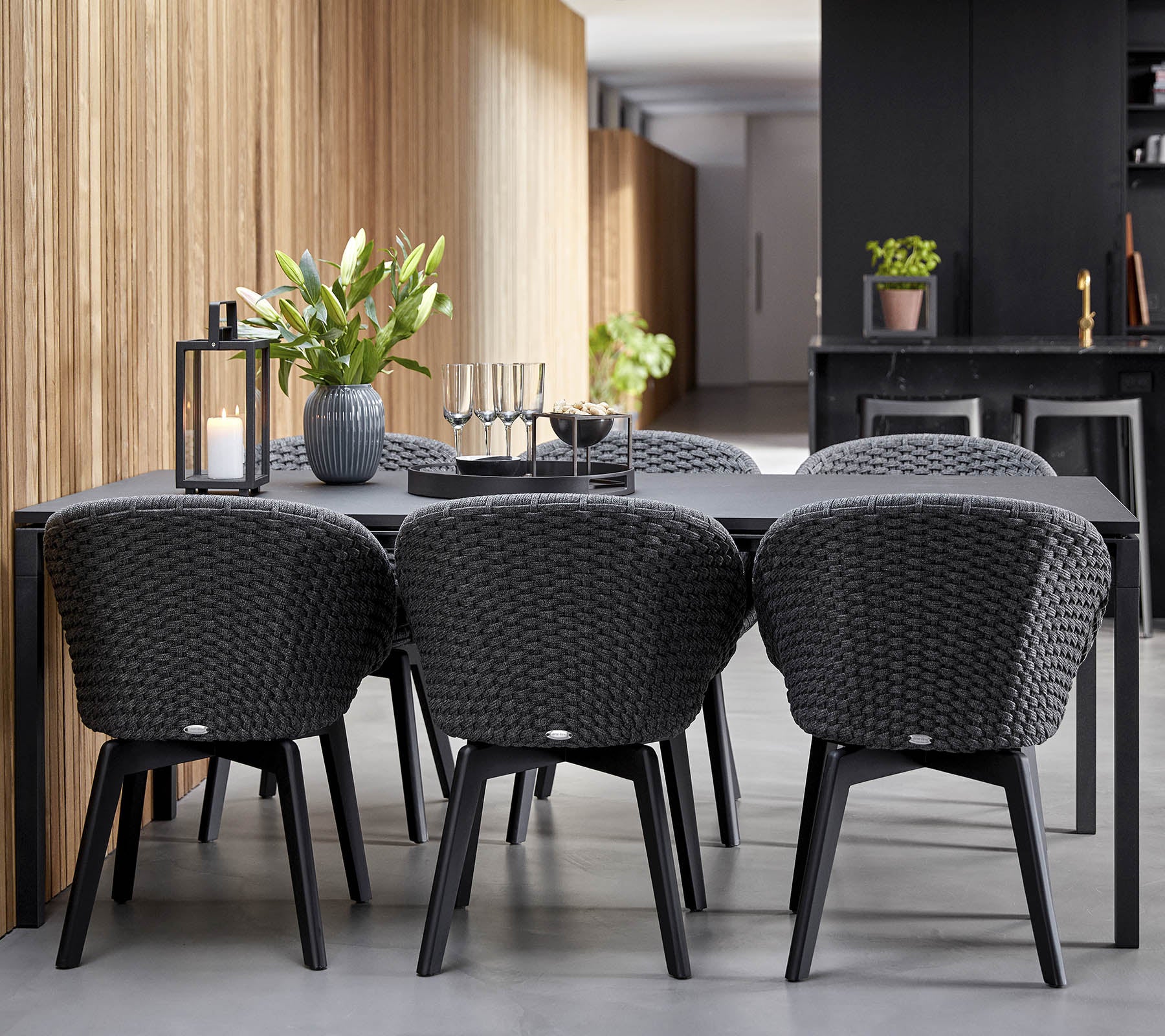 Una configuración de comedor moderna con una elegante mesa negra rodeada de sillas trenzadas con estilo, complementada con plantas decorativas y velas.
