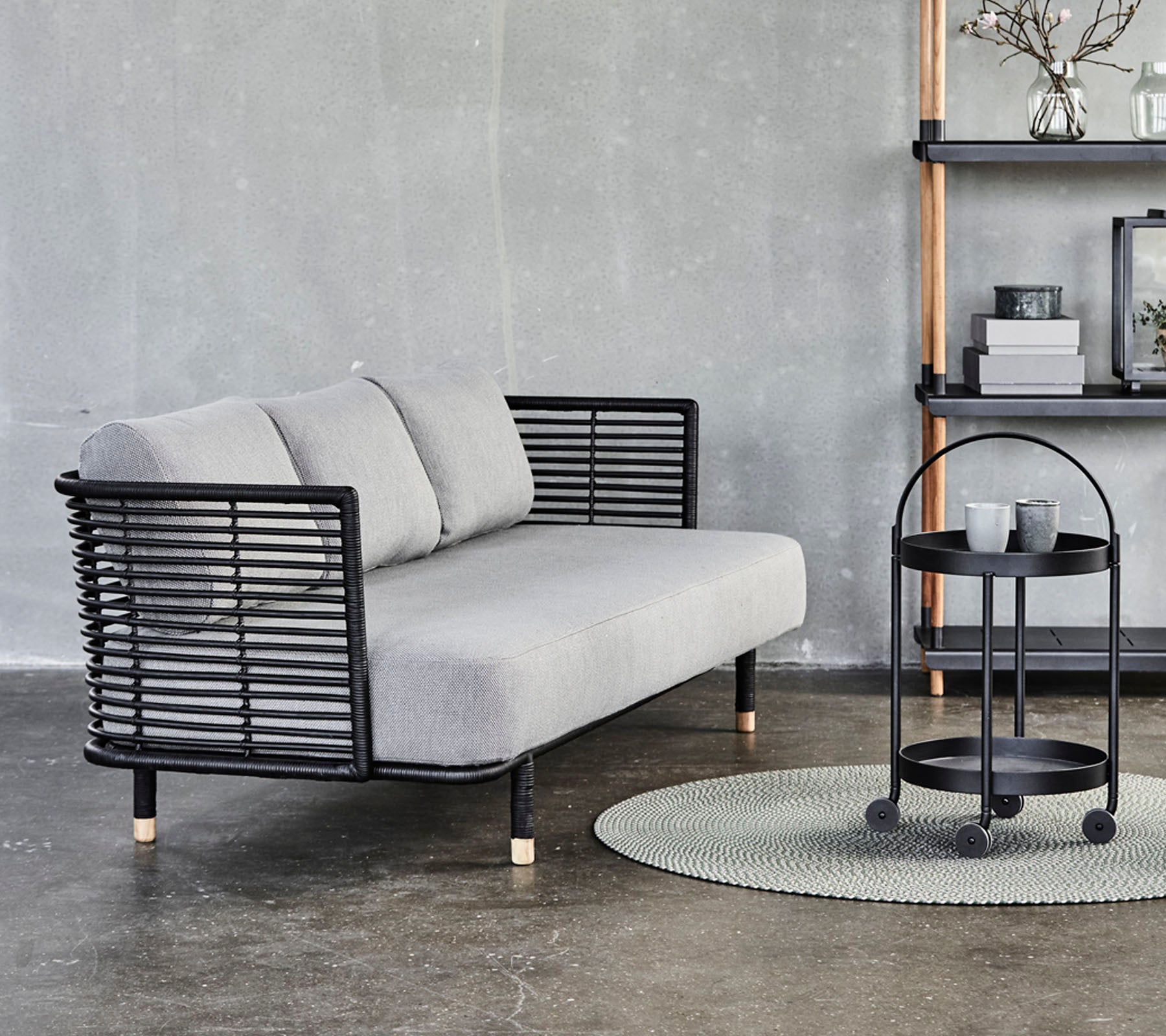 Sofá gris elegante con acentos de metal negro y cojines, acompañado de una mesa lateral redonda sobre una alfombra texturizada contra un fondo neutro.
