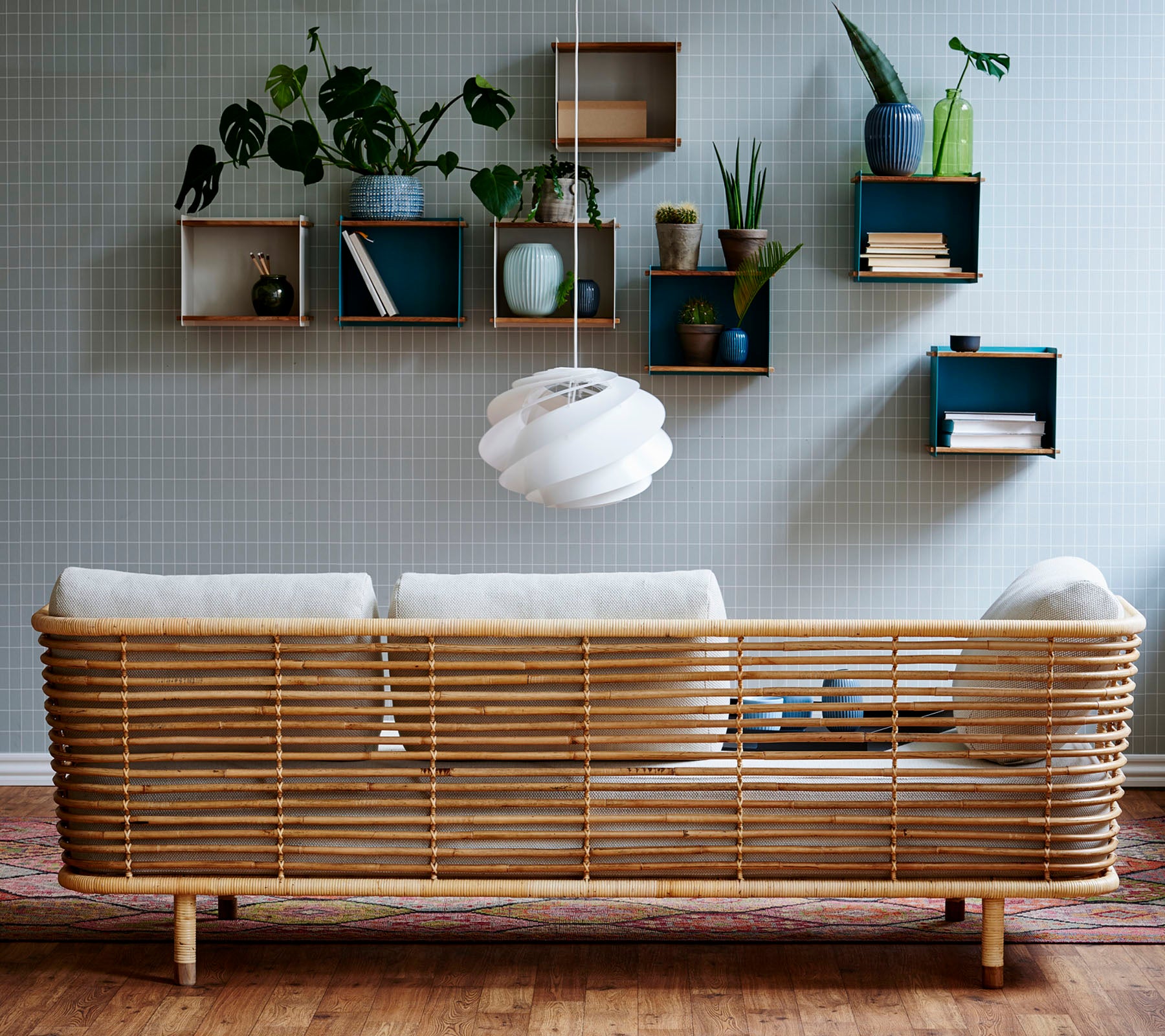 Un sofá de ratán elegante con cojines claros contra una pared texturizada, complementado por estanterías montadas en la pared que exhiben plantas y elementos decorativos.
