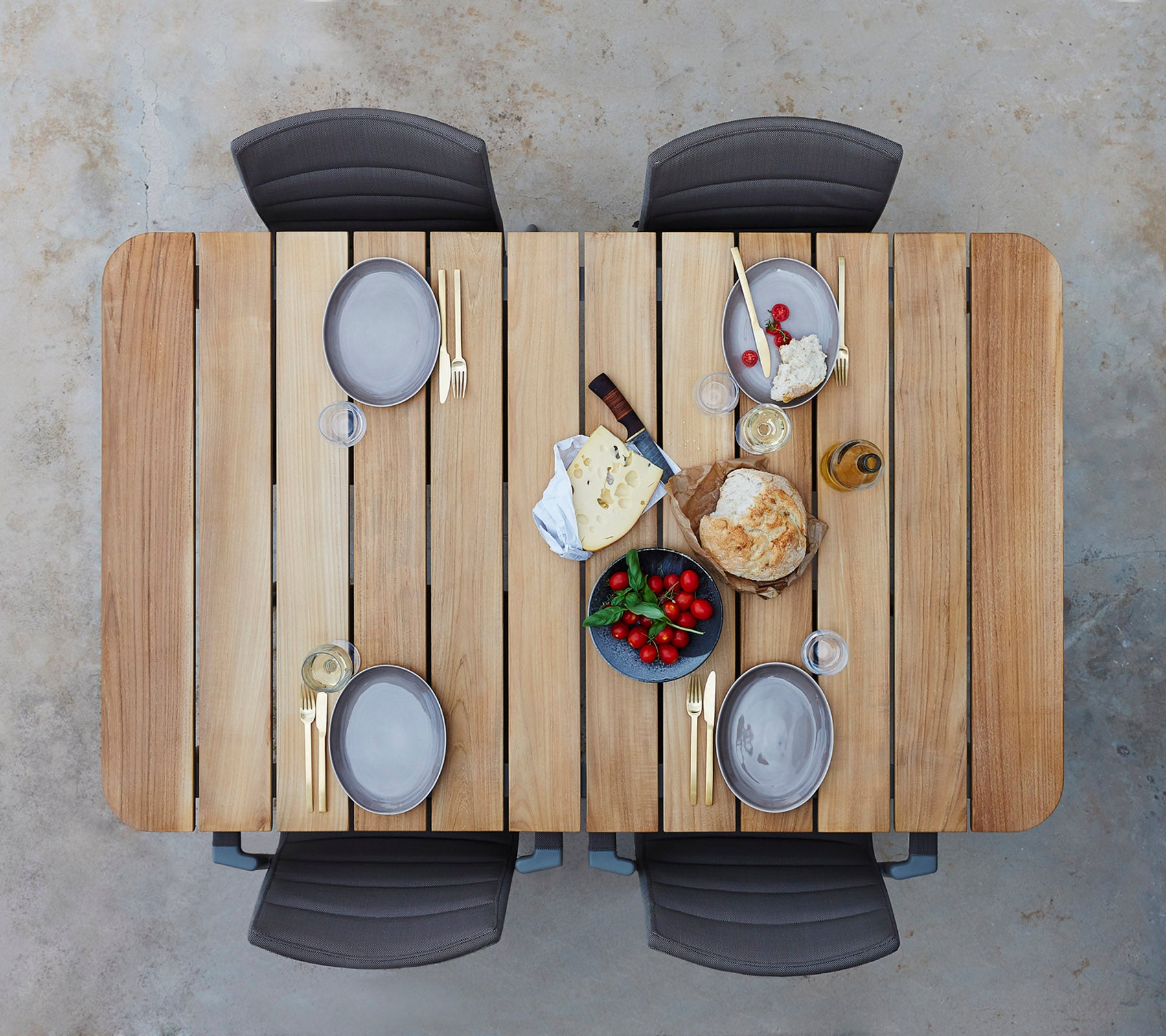 Una mesa de madera servida con platos, vasos y una variedad de quesos, frutas y pan, rodeada de cuatro sillas.