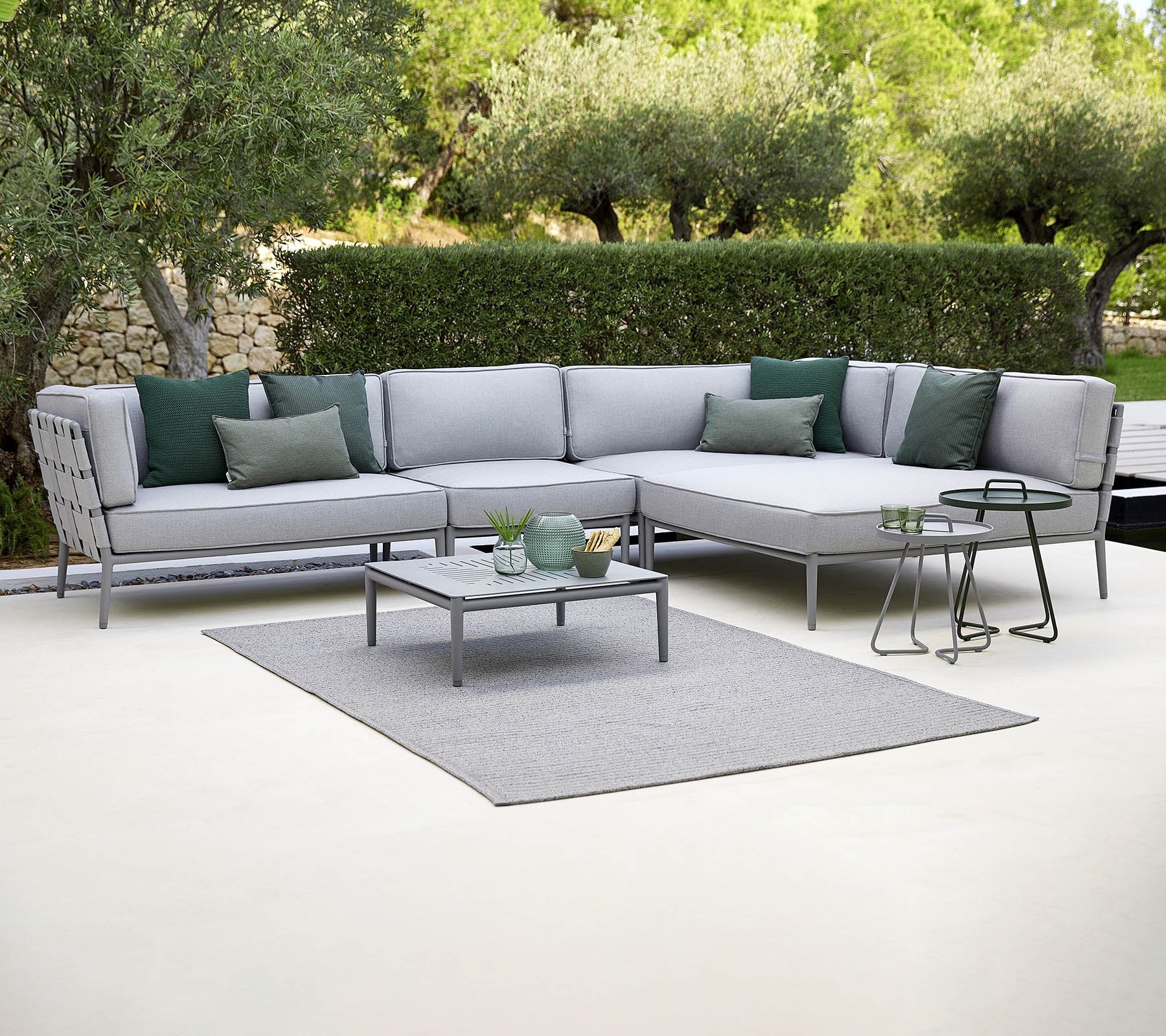 Sofá seccional exterior elegante con cojines verdes, una mesa de café y una alfombra, con un fondo de vegetación.