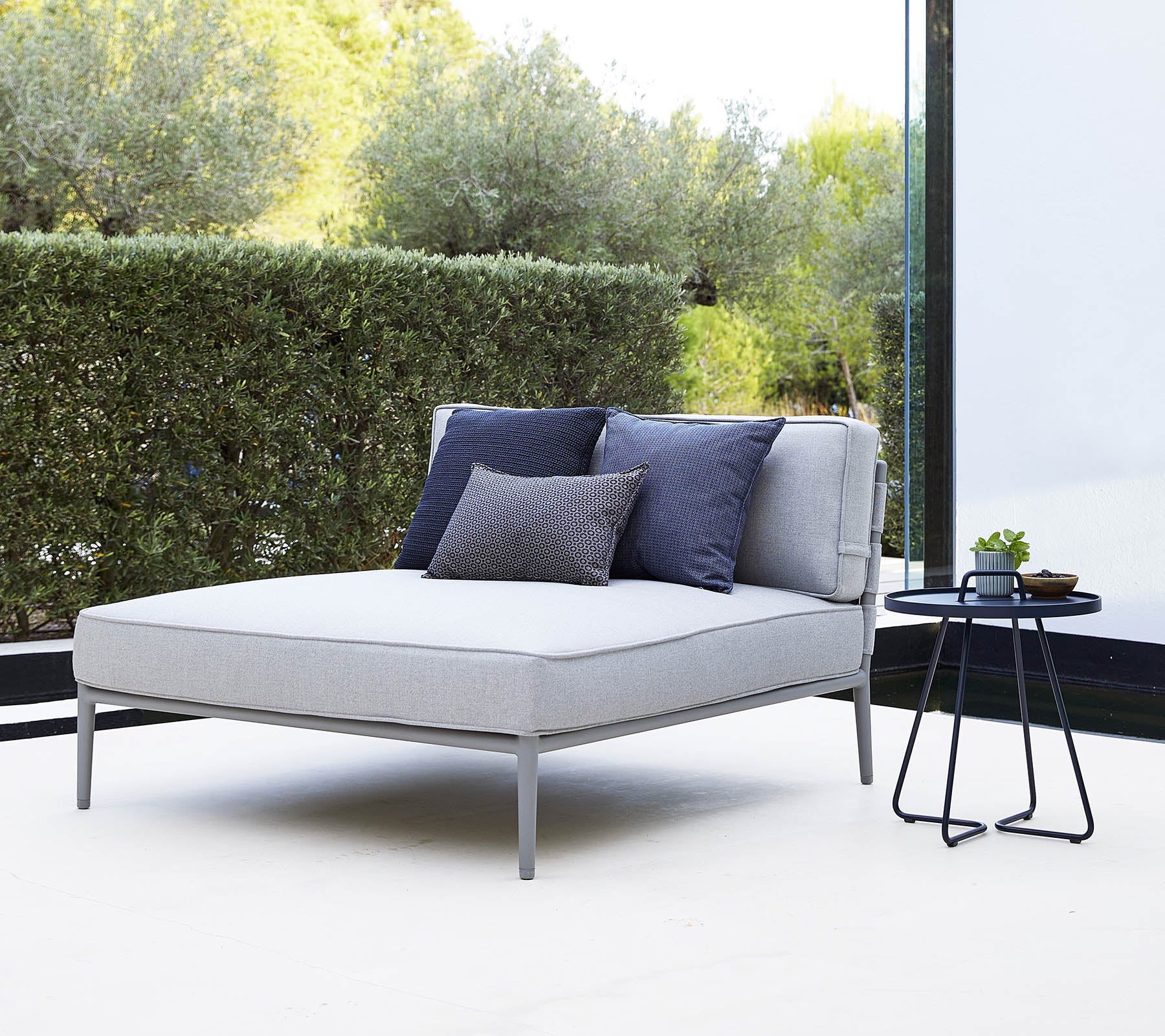 Sofá lounge gris elegante con cojines azul marino al lado de una pequeña mesa auxiliar negra en un entorno sereno.